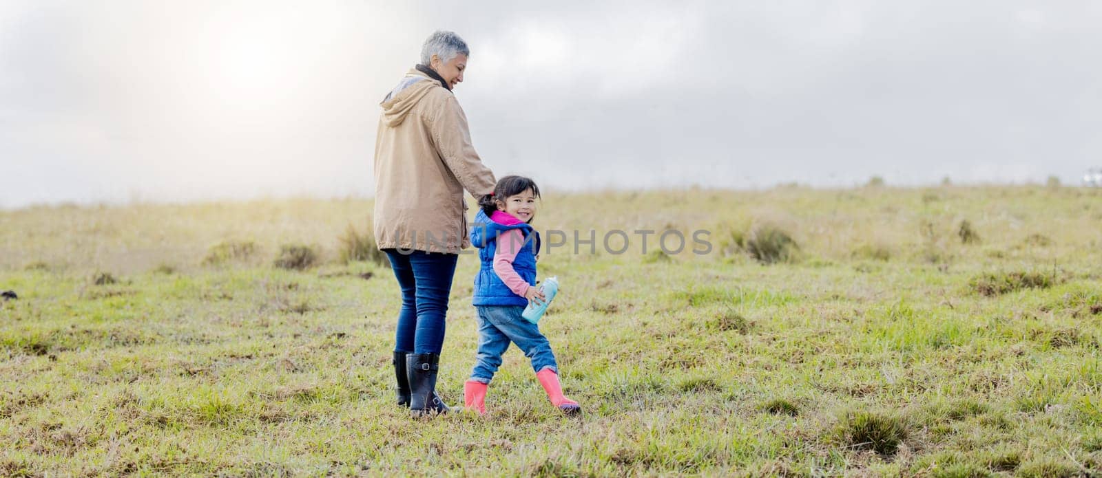 Grandmother, walking girl and nature park of a kid with senior woman in the countryside. Outdoor field, grass and elderly female with child on a family adventure on vacation with happiness and fun.