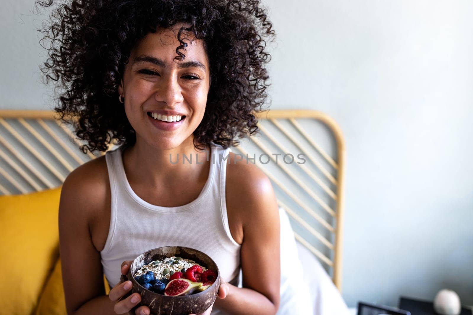 Happy young multiracial woman smiling looking at camera showing bowl of healthy breakfast oats with fruit sitting on bed. Breakfast in bed. Healthy lifestyle concept.