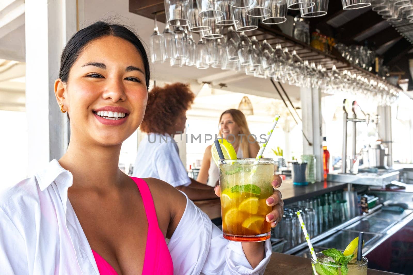 Happy young latina woman showing mojito cocktail looking at camera at a beach bar in summertime. by Hoverstock