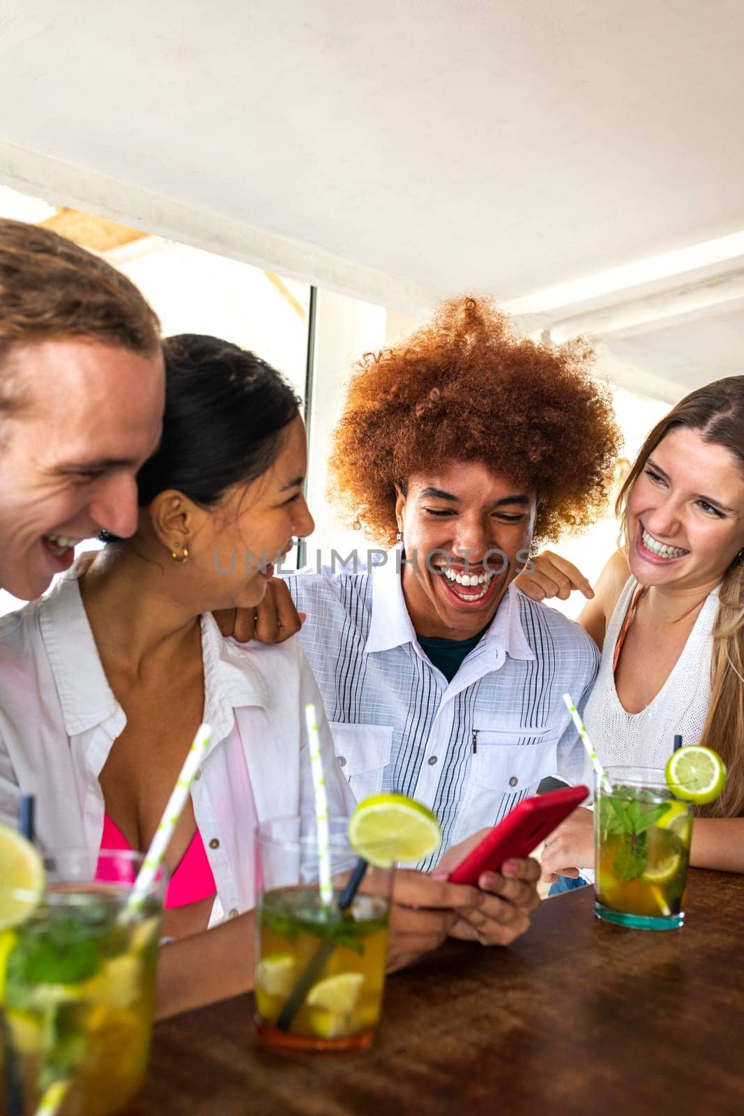 Young Latina and her happy multiracial friends looking at mobile phone in a beach bar having mojito cocktails. Vertical image. Technology and leisure activity.