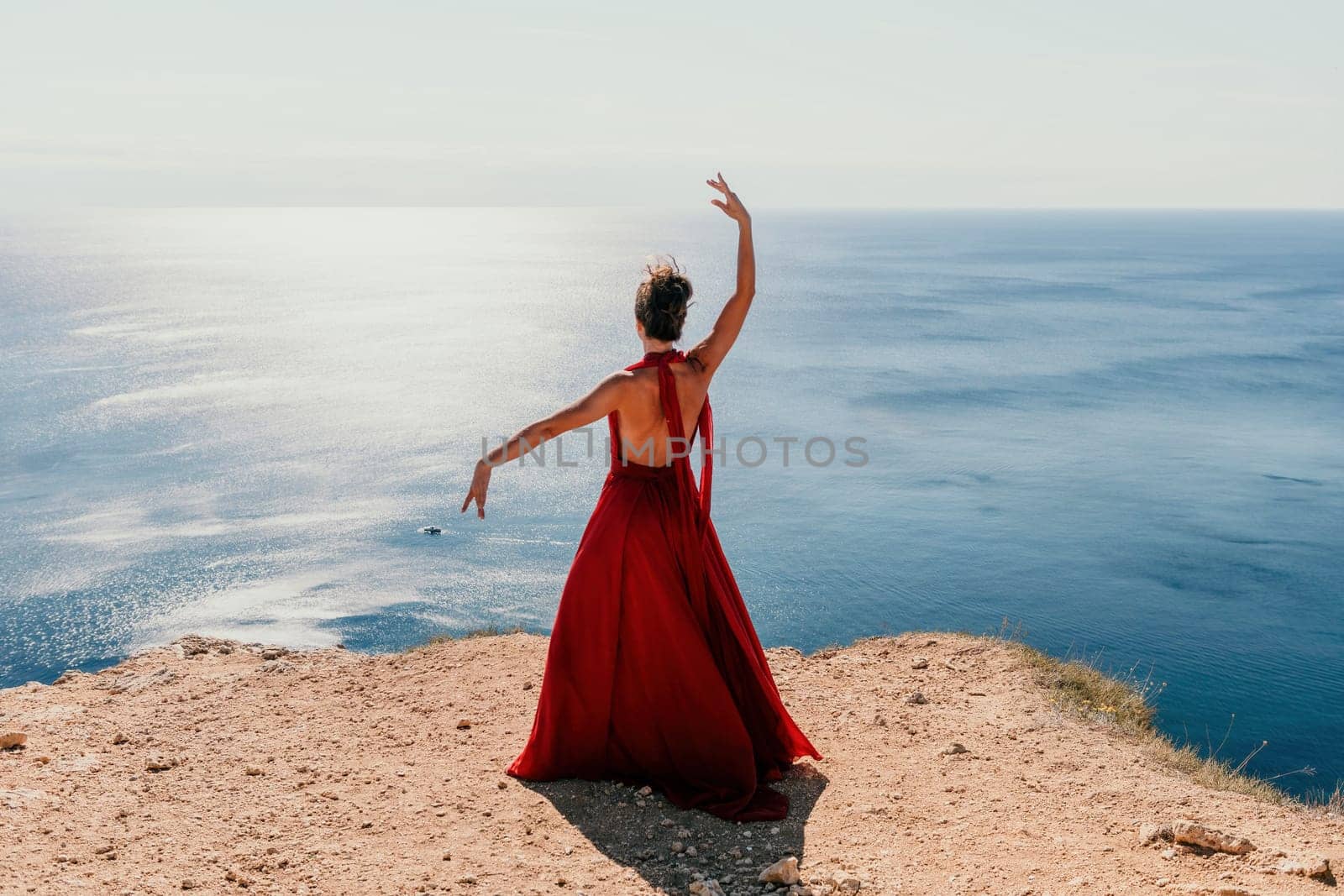 Woman in red dress on sea. Side view a Young beautiful sensual woman in a red long dress posing on a rock high above the sea on sunset. Girl on the nature on blue sky background. Fashion photo. by panophotograph