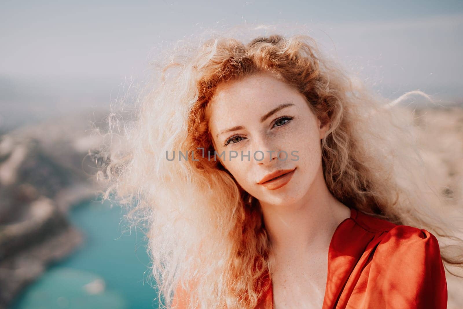 Close up portrait of curly redhead young caucasian woman with freckles looking at camera and smiling. Cute woman portrait in a pink long dress posing on a volcanic rock high above the sea at sunset by panophotograph