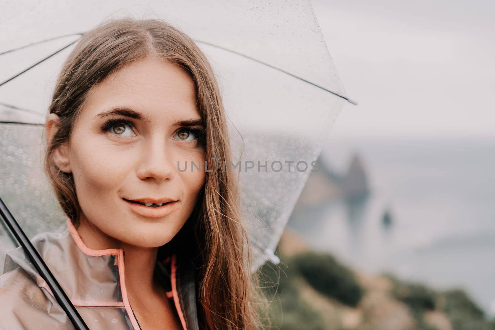 Woman rain umbrella. Happy woman portrait wearing a raincoat with transparent umbrella outdoors on rainy day in park near sea. Girl on the nature on rainy overcast day. by panophotograph