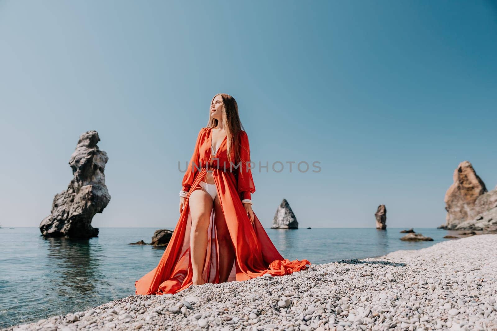 Woman travel sea. Happy tourist in red dress enjoy taking picture outdoors for memories. Woman traveler posing on the rock at sea bay surrounded by volcanic mountains, sharing travel adventure journey by panophotograph