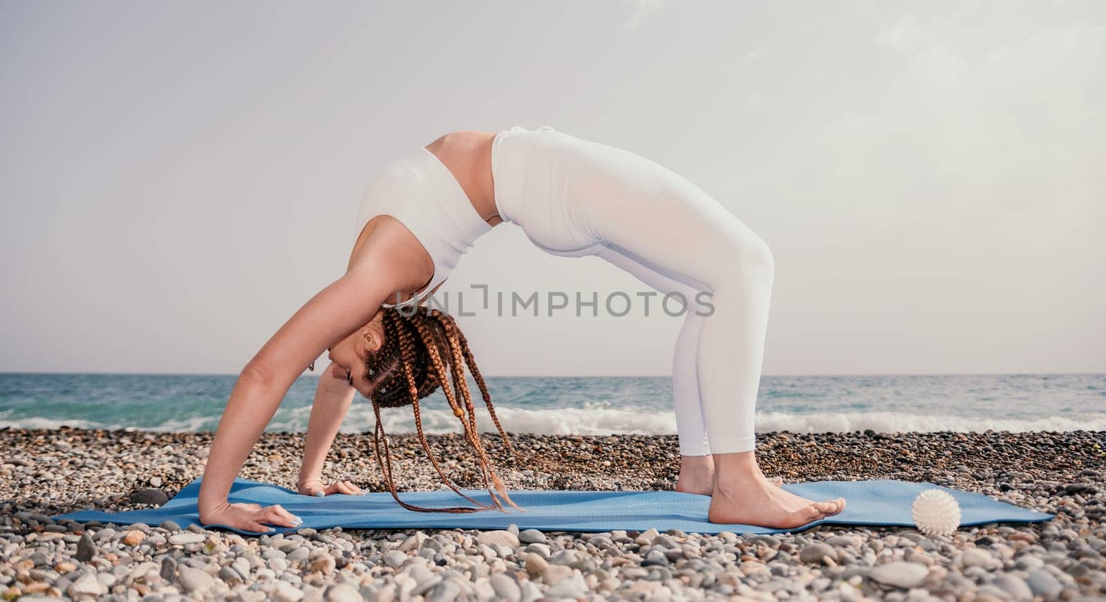 Woman sea yoga. Well looking middle aged woman with braids dreadlocks in white leggings and tops doing stretching pilates on yoga mat near sea. Female fitness yoga routine concept. Healthy lifestyle. by panophotograph