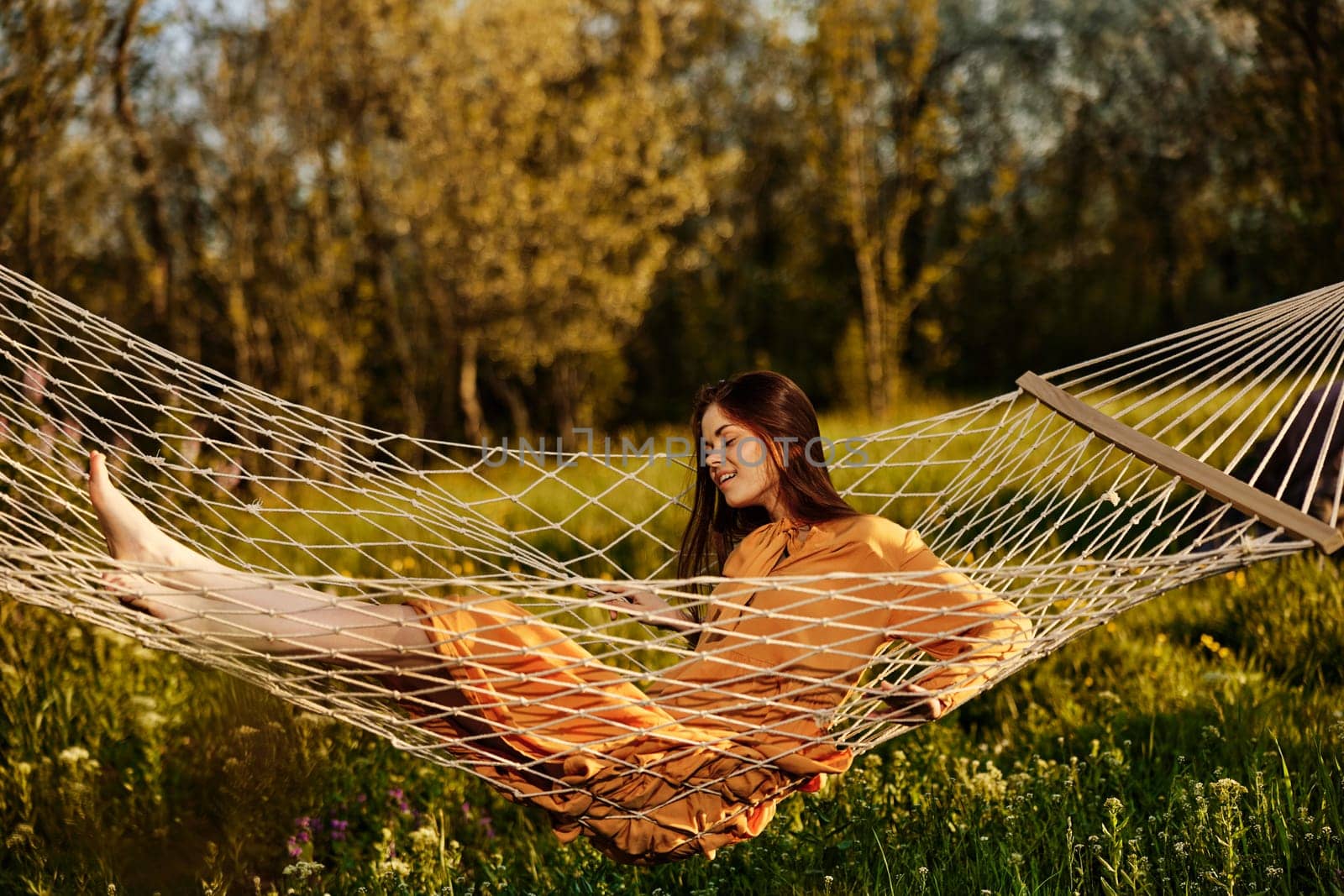 horizontal photo of a beautiful, red-haired woman lying in a hammock enjoying a rest in a long orange dress, on a warm summer day, smiling happily by Vichizh