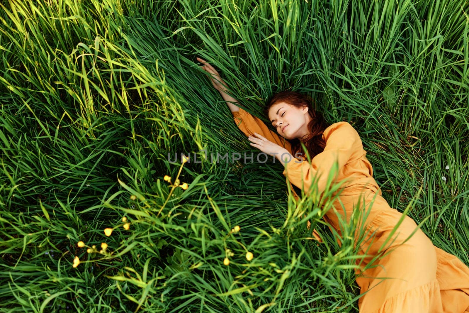 a relaxed woman enjoys summer lying in the tall green grass in a long orange dress stretched out stretching her arm forward. High quality photo