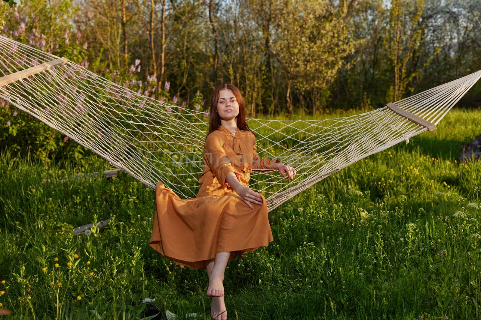 horizontal photo of a beautiful, red-haired woman sitting on the edge in a hammock enjoying a rest in a long orange dress, on a warm summer day, happily looking into the camera. High quality photo