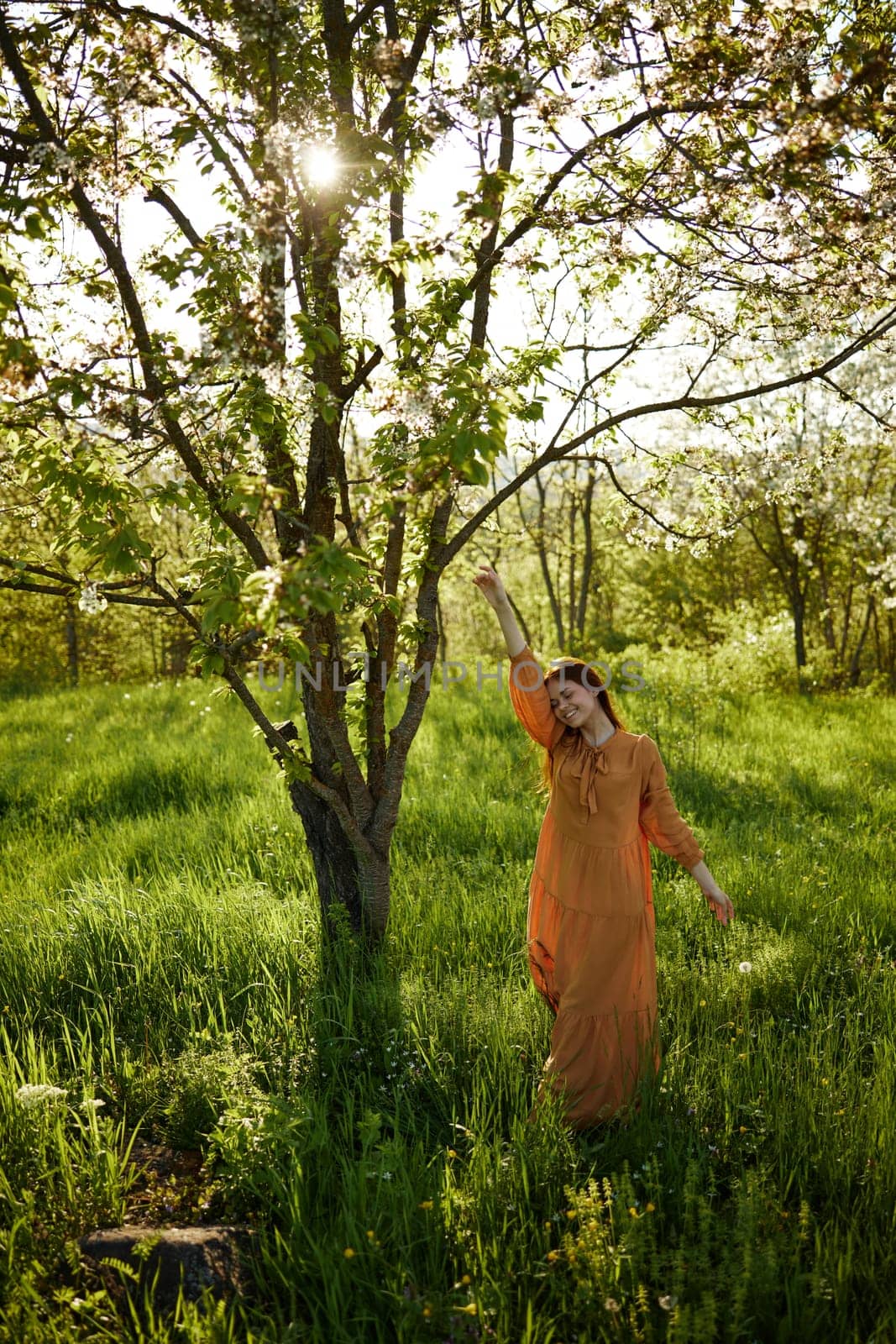 a beautiful, joyful woman stands in a long orange dress, in the countryside, near a tree blooming with white flowers, during sunset, illuminated from the back and reaching for the branches of the tree by Vichizh