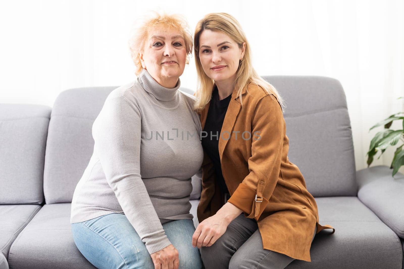 Beautiful senior mom and her adult daughter are hugging and smiling while sitting on couch at home.
