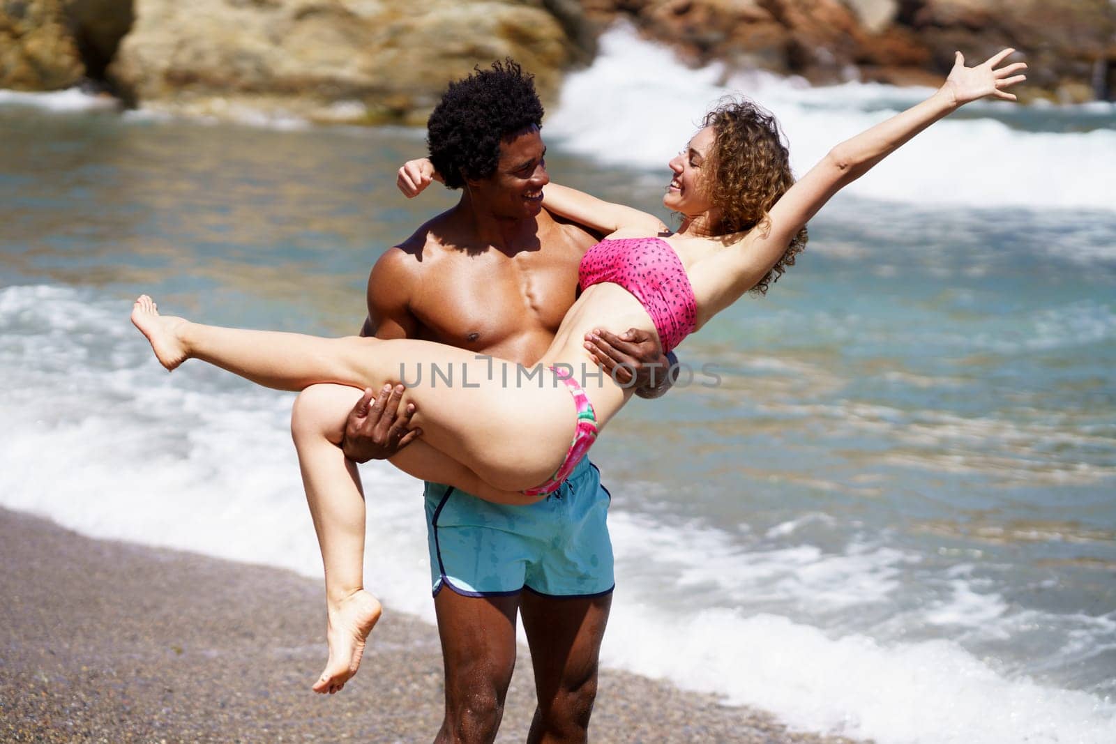 Smiling young multiracial couple enjoying near waving sea, and looking at each other while black male carrying girlfriend with stretched hand and standing on beach in daylight