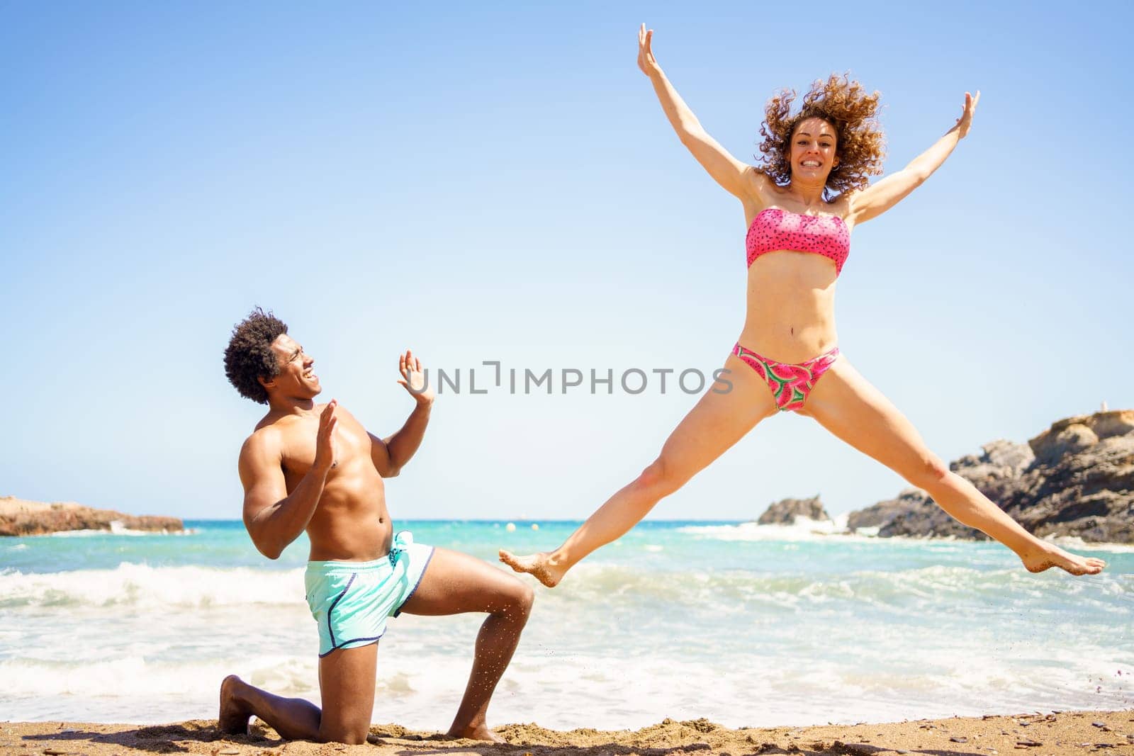 Full body of happy young woman with curly hair jumping on sand with outstretched arms and legs near kneeling African American boyfriend