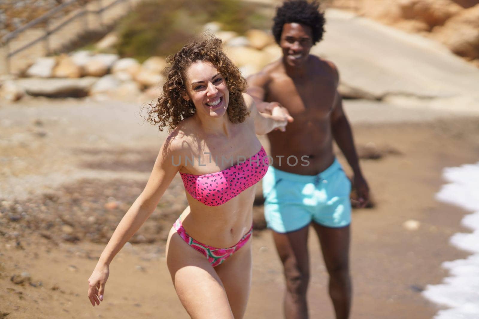 Happy young multiethnic couple in swimwear holding hands while running on sandy seashore during summer sunny day having fun together