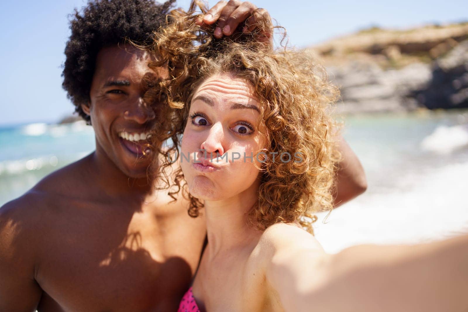 Cheerful young woman with puffed out cheeks taking selfie with black boyfriend touching curly hair of girlfriend while having fun on beach