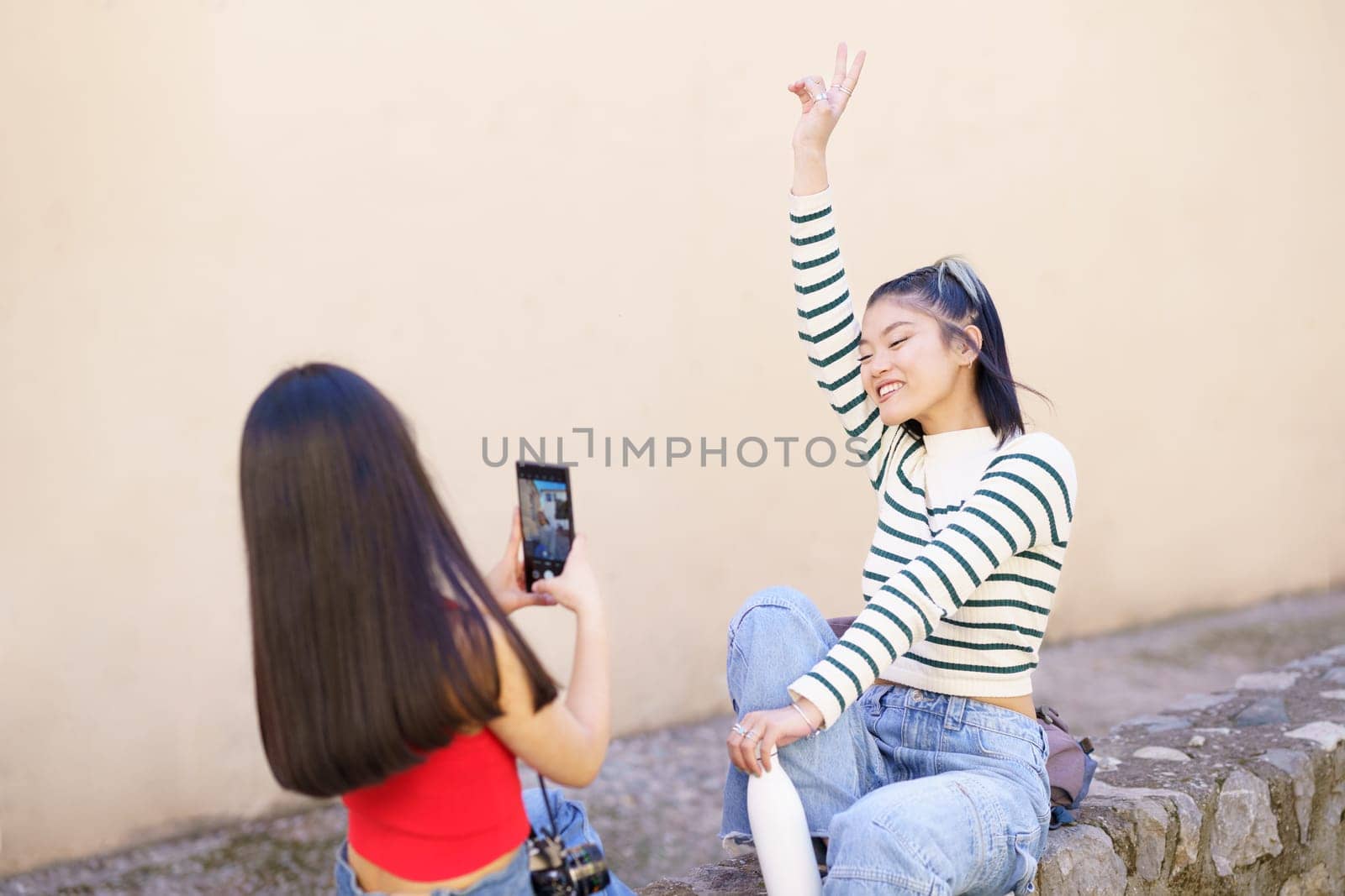 Happy young Asian women tourists taking picture on smartphone wile sitting on stone border on town street during vacation together