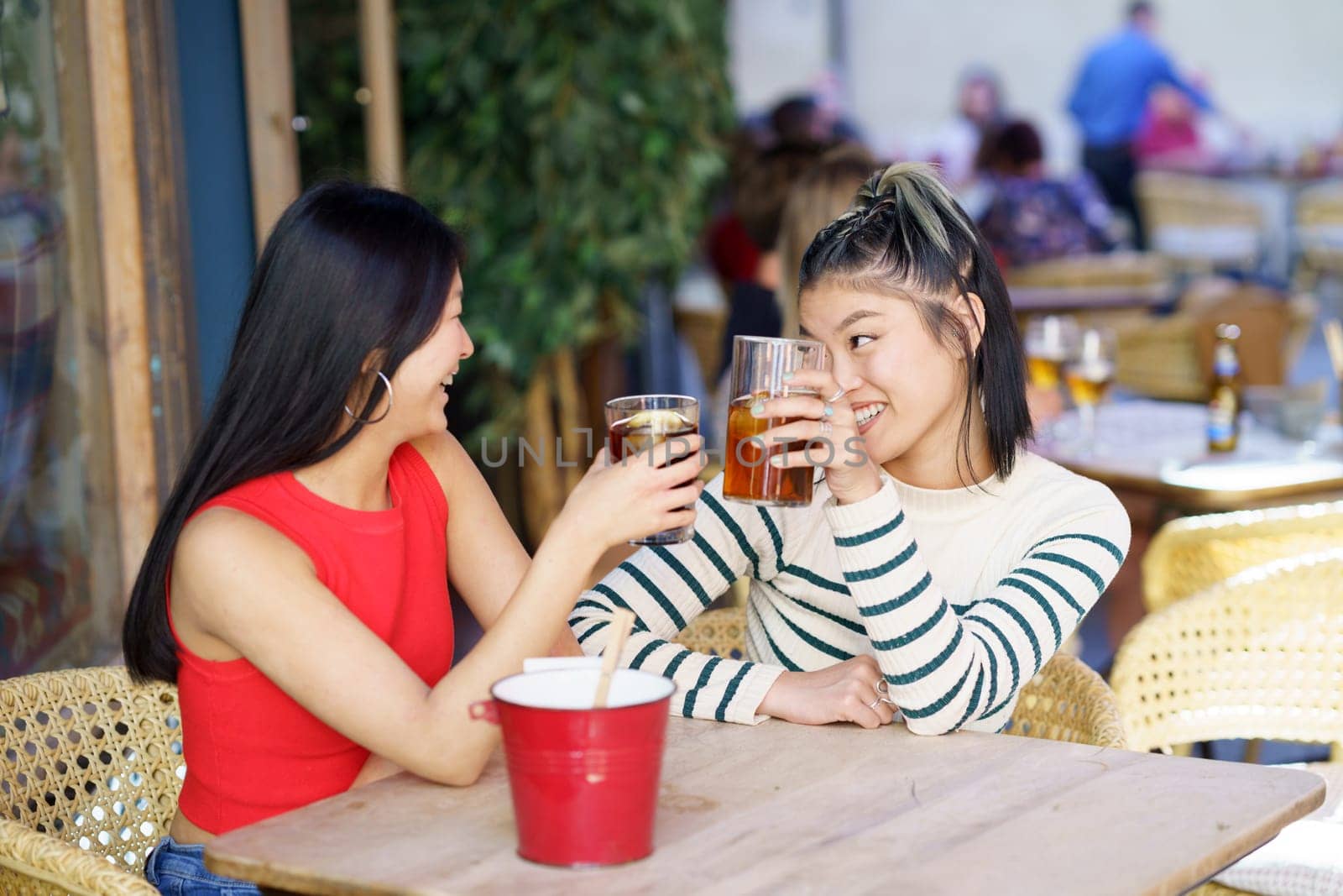 Positive young Asian female friends in casual clothes sitting together at wooden table, and clinking glasses of fresh beverages while looking at each other during meeting in cafe