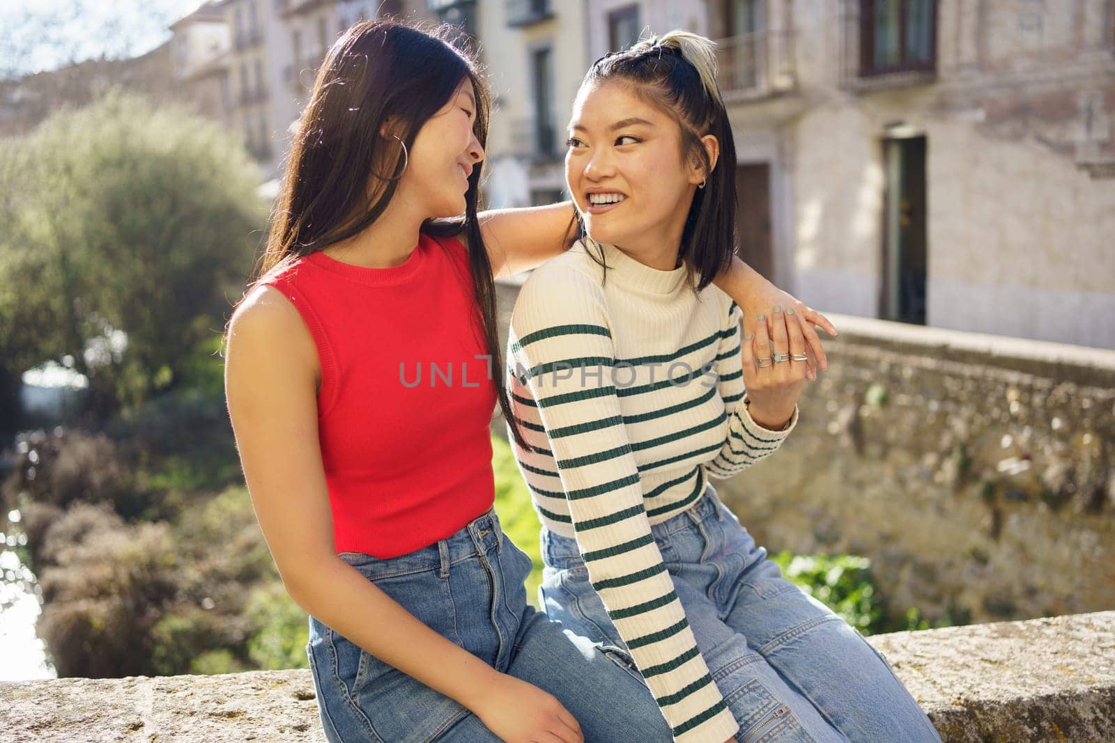 Happy Asian girlfriends sitting together on street and looking at each other by javiindy