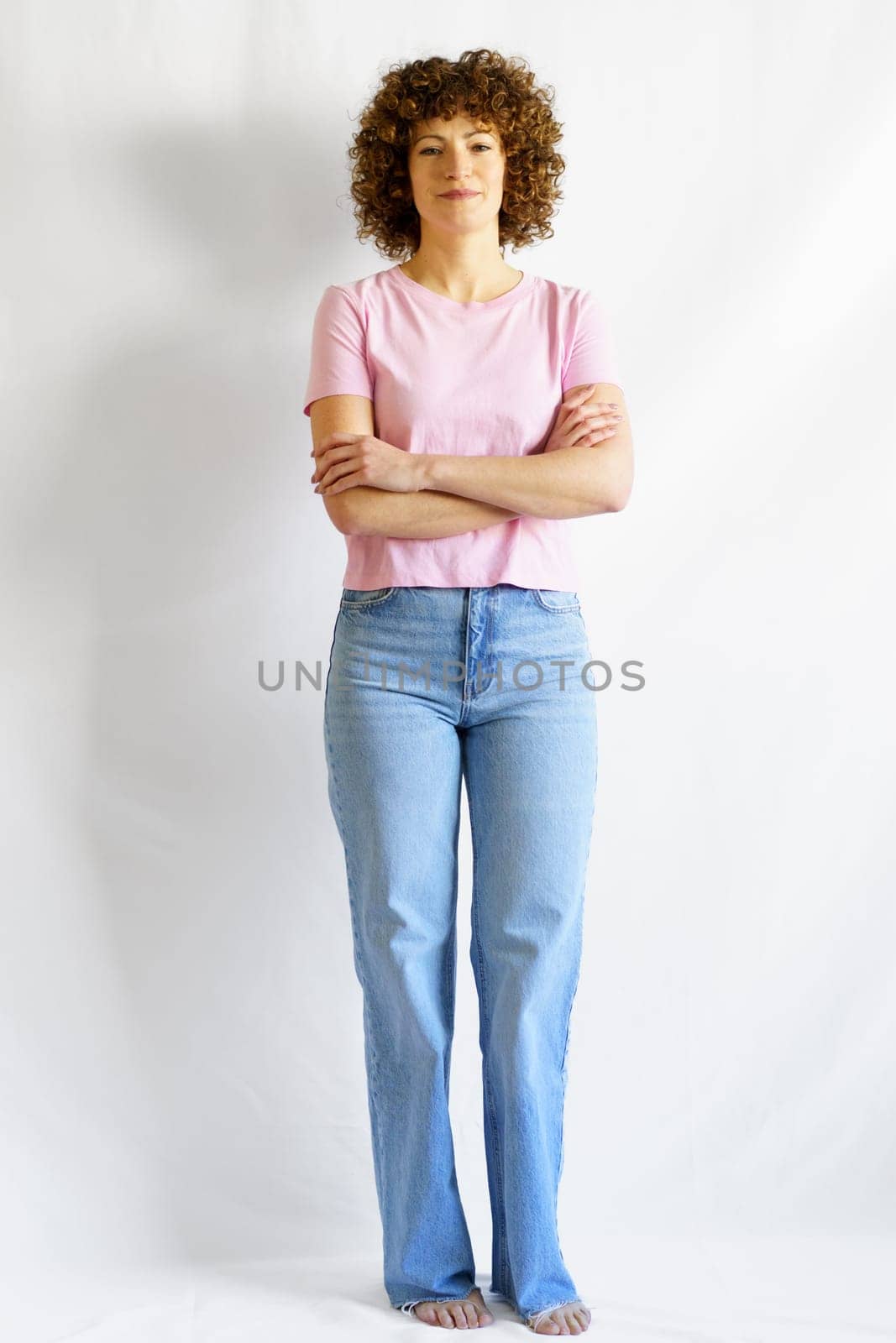 Full body of curly haired female in jeans and pink basic t-shirt leaning on white wall with hands crossed looking at camera