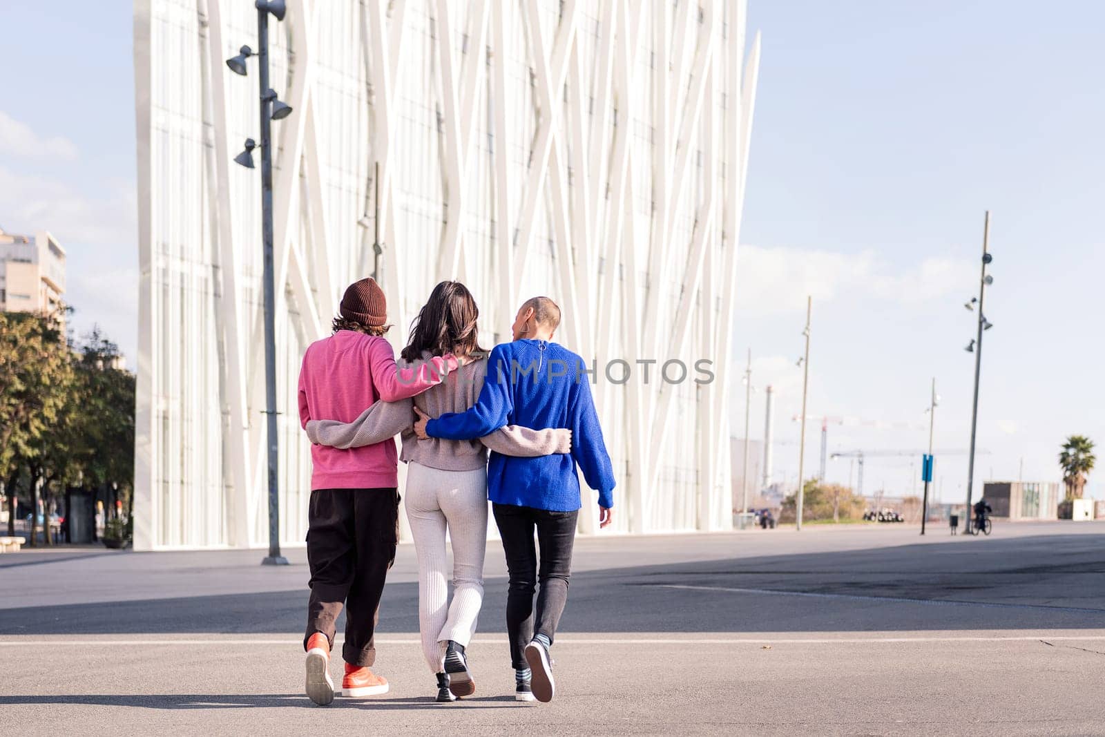 rear view of three unrecognizable friends, one man and two women, embracing while walking on a sunny day, concept of friendship and urban lifestyle