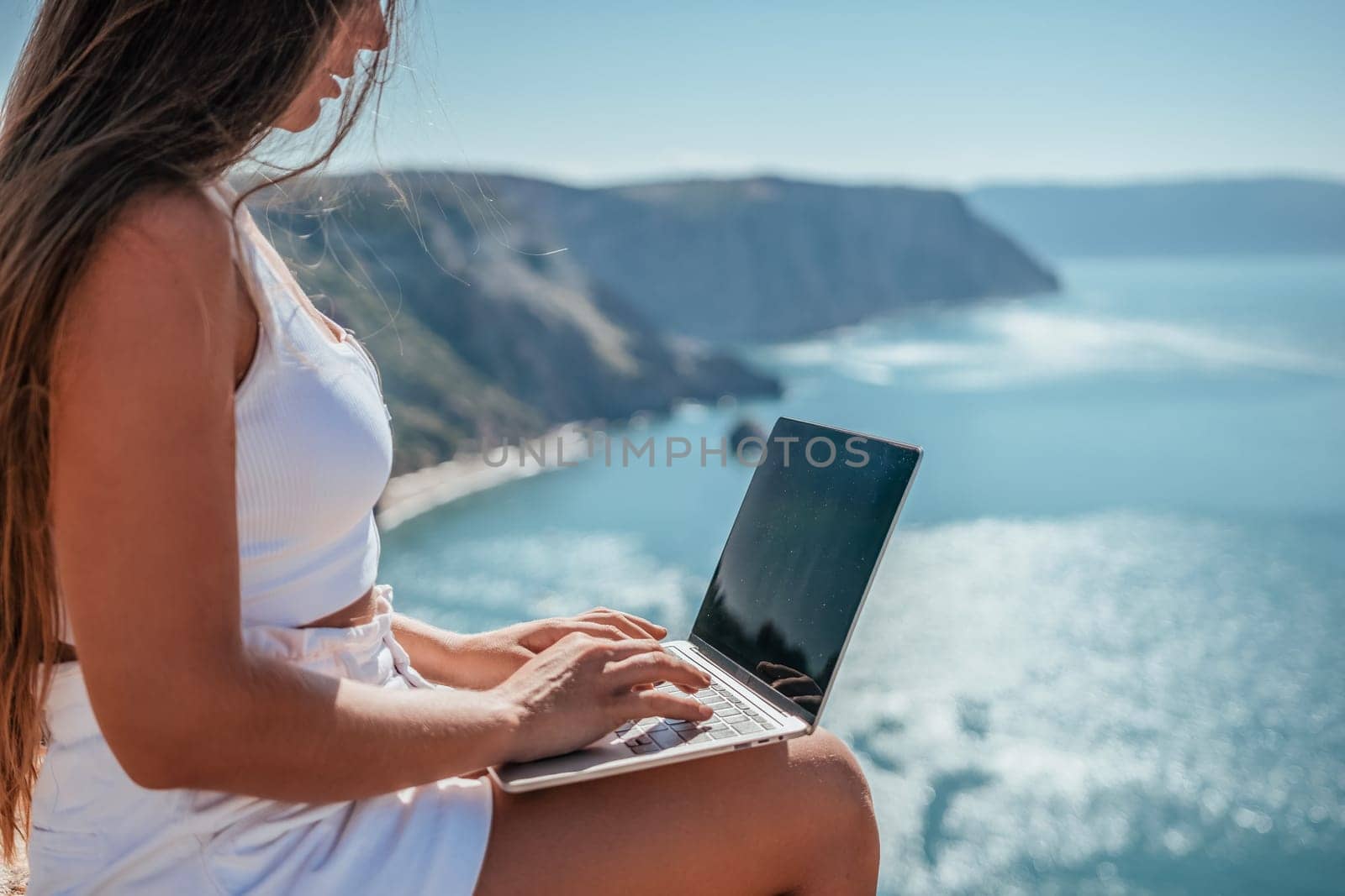 Successful business woman in yellow hat working on laptop by the sea. Pretty lady typing on computer at summer day outdoors. Freelance, travel and holidays concept.