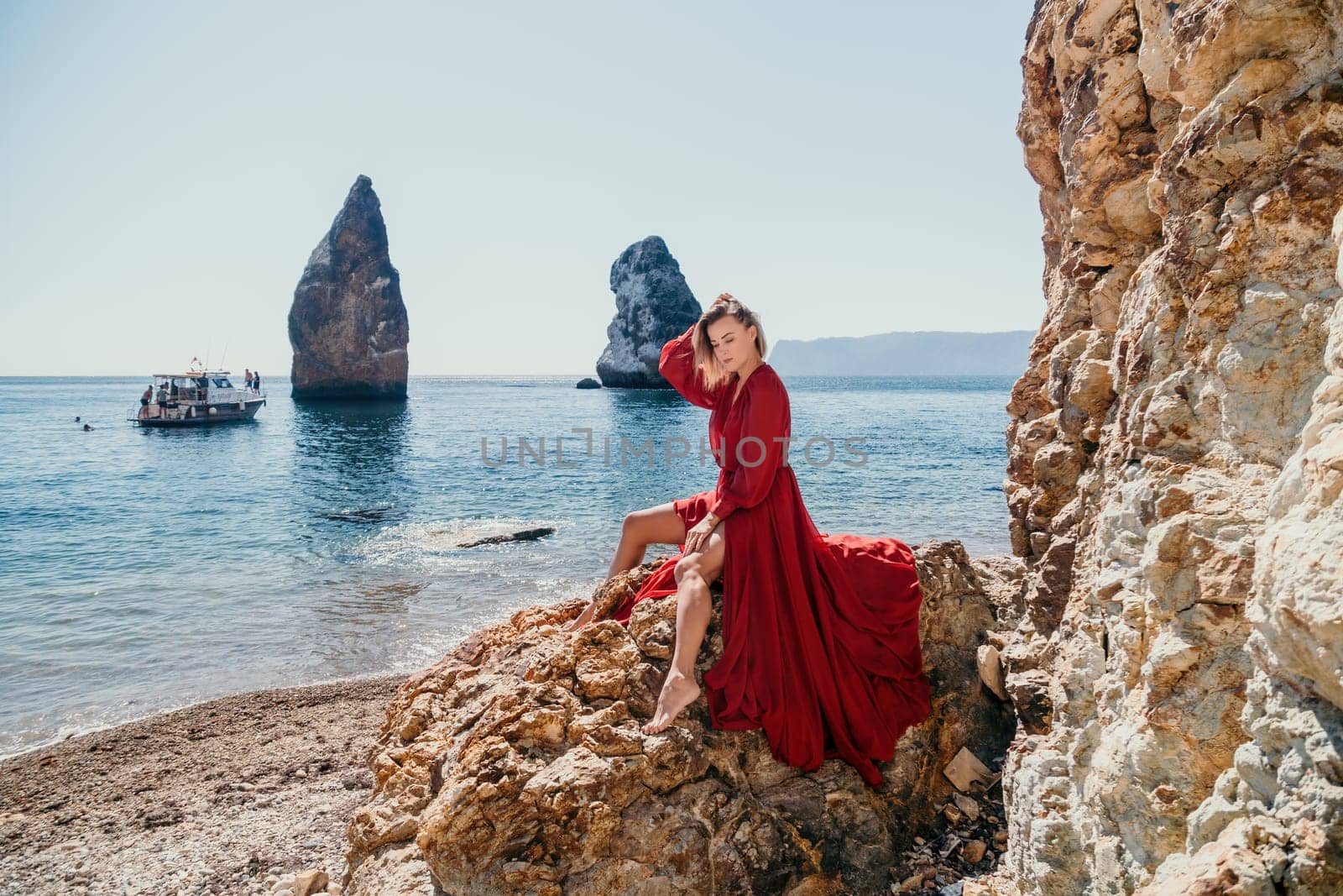 Woman summer travel sea. Happy tourist in long red dress enjoy taking picture outdoors for memories. Woman traveler posing on beach at sea surrounded by volcanic mountains, sharing travel adventure by panophotograph