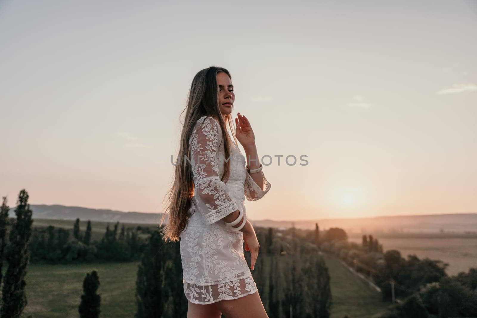 Happy woman in white boho dress on sunset in mountains. Romantic woman with long hair standing with her back on the sunset in nature in summer with open hands. Silhouette. Nature. Sunset. by panophotograph