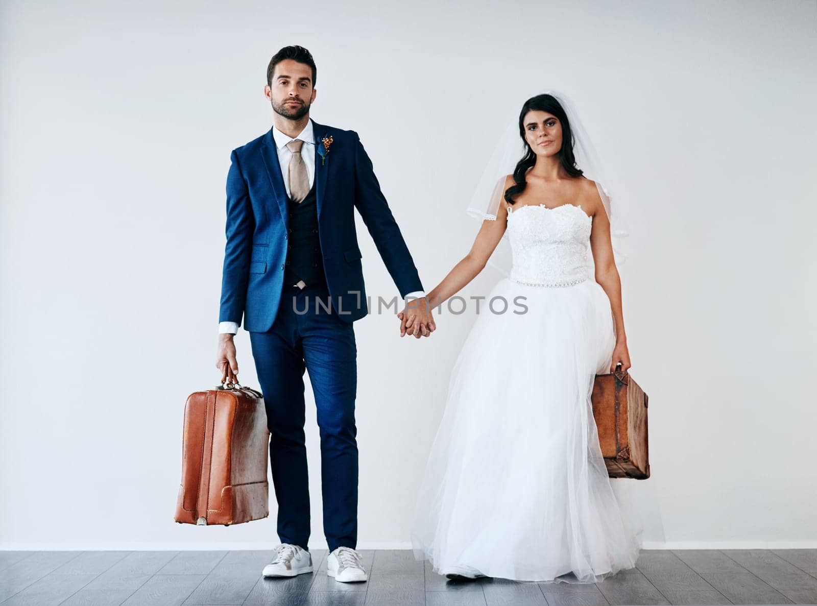 We have our travel shoes on. Studio shot of a newly married couple holding hand and carrying bags while standing against a gray background