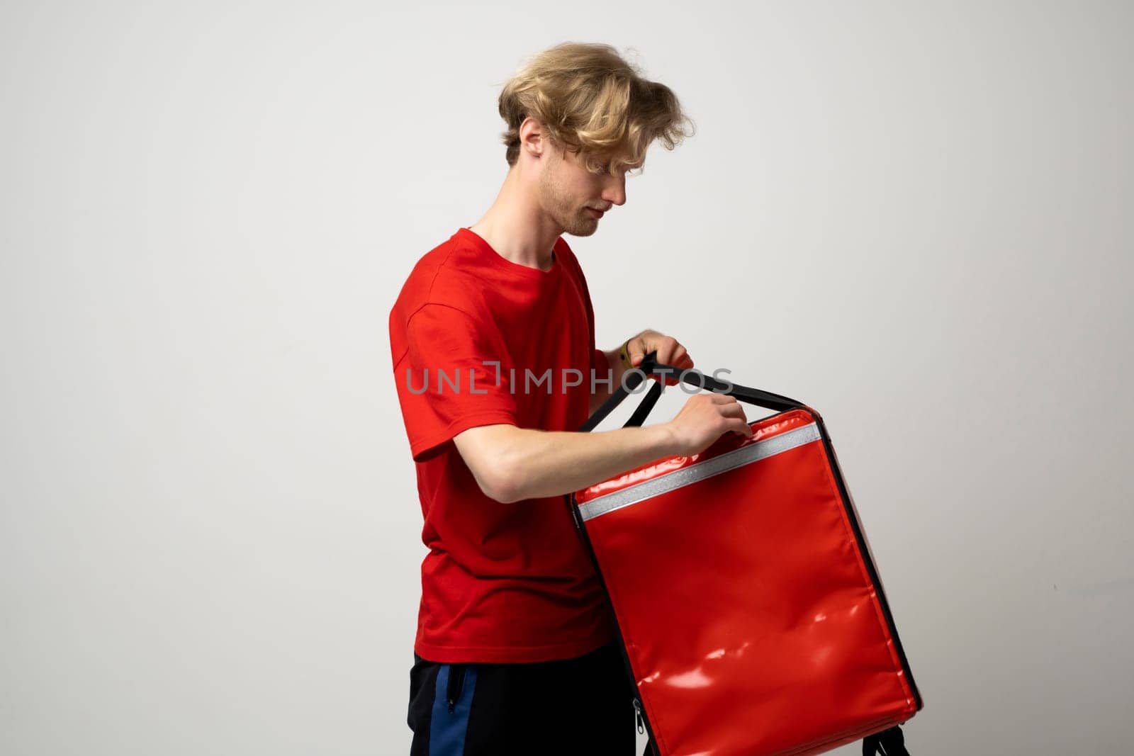 Young courier opening thermal bag on white background. Food delivery service. Delivery guy in a red t-shirt uniform work as courier and holds red thermal food backpack. Service concept. by vovsht