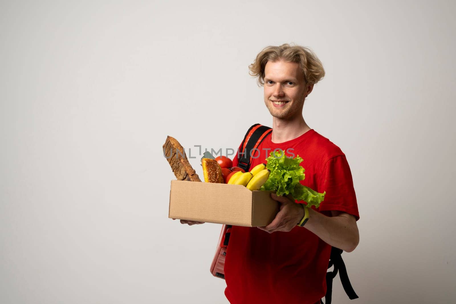 Courier man in red uniform with a thermal bag backpack hold craft paper bag takeaway with food products isolated on white background studio. Delivery service from shop restaurant to home. by vovsht