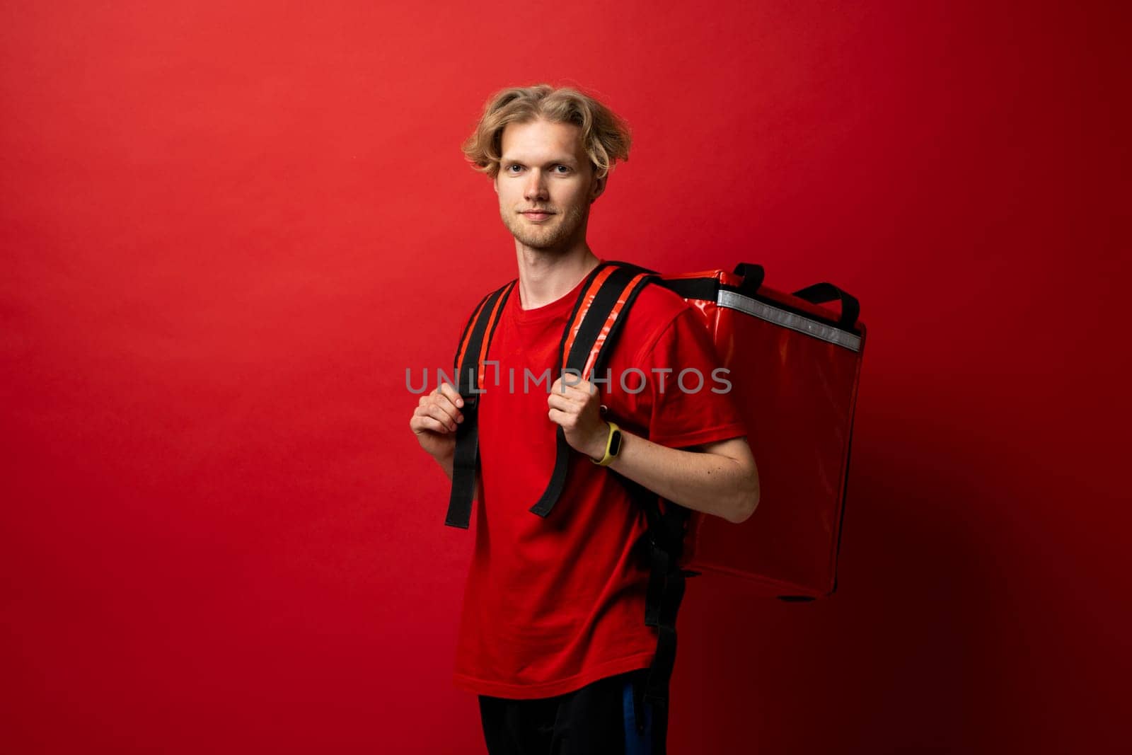 Portrait of happy smiling delivery man in a red t-shirt with thermal insulated bag on a shoulder looking in a camera. Food delivery service. by vovsht