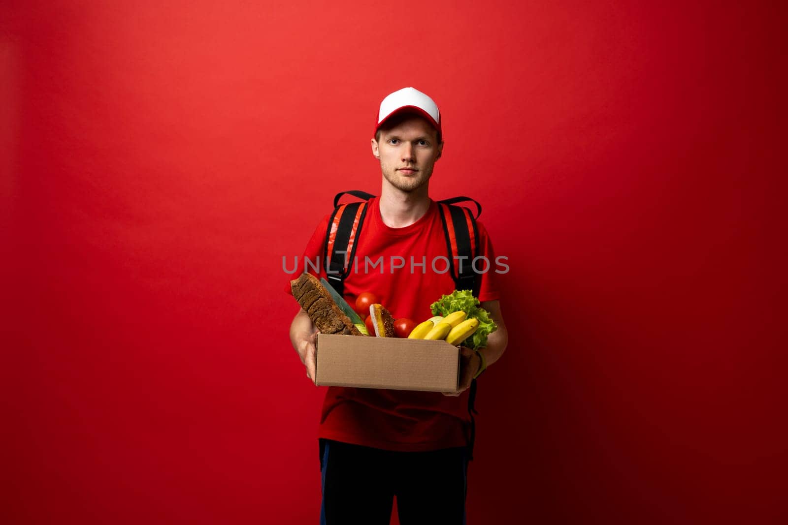 Delivery Concept. Portrait of pleased delivery man in red uniform smiling while carrying paper bag with food products isolated over red background. Food delivery service. by vovsht