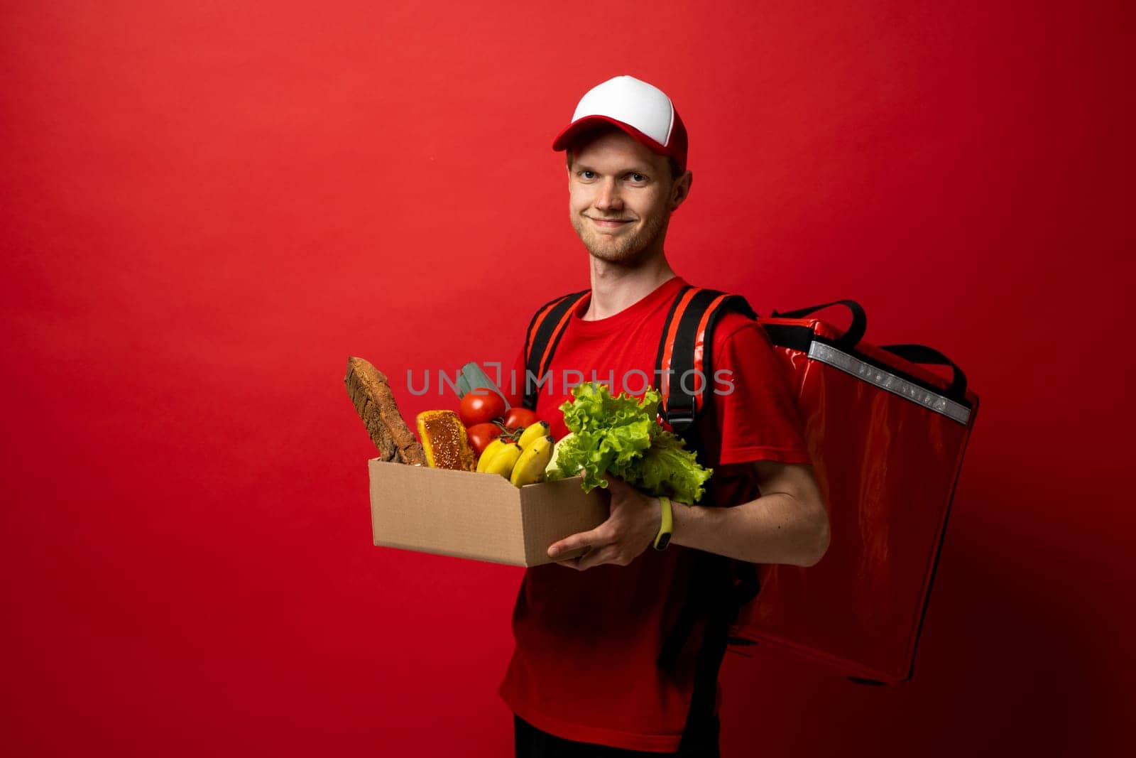 Delivery male employee in a red uniform holds paper cardboard package with organic groceries, fruits on a red background. Products delivery from shop or restaurant to home. Courier service