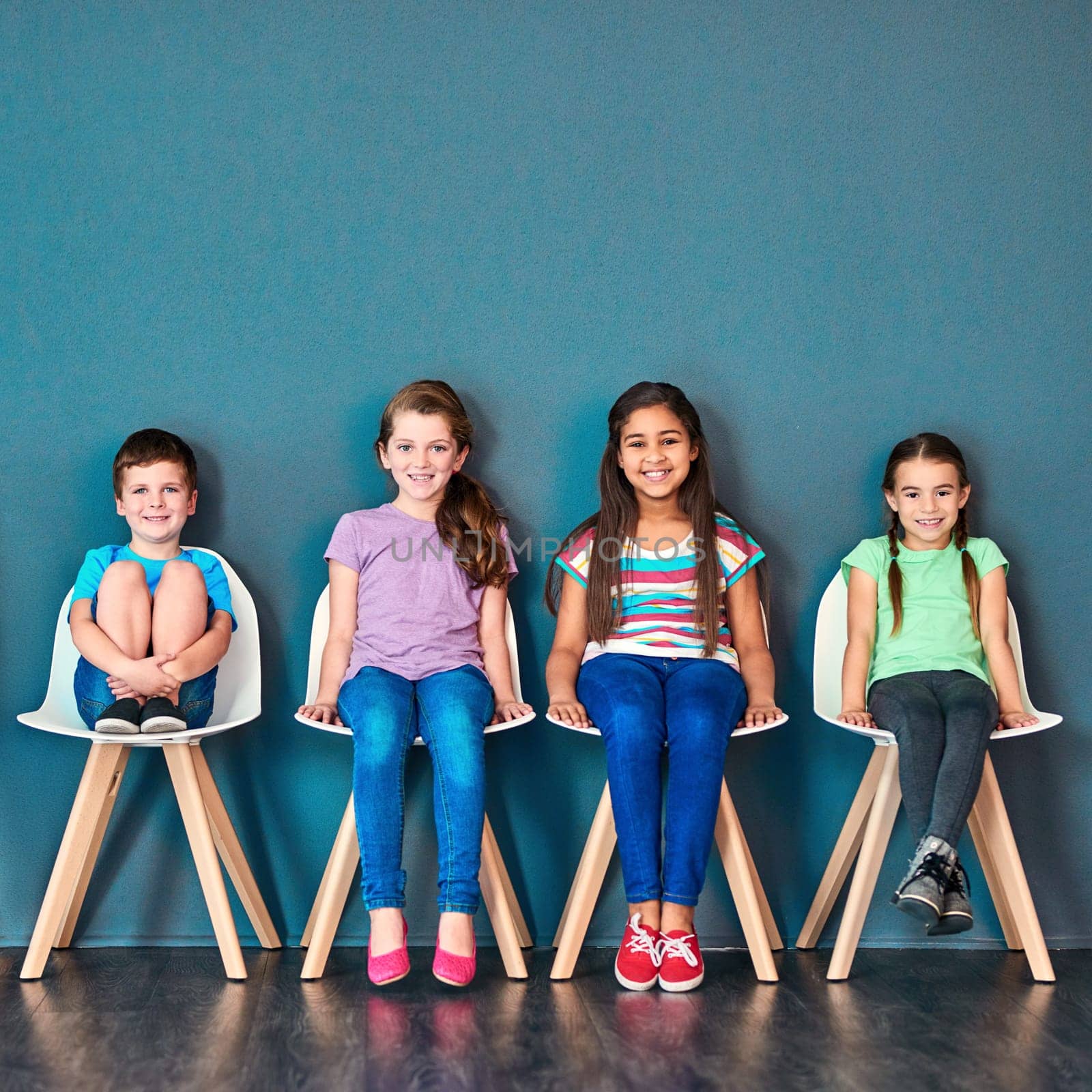 Its our turn now. Studio portrait of a diverse group of kids sitting on chairs in a line against a blue background. by YuriArcurs