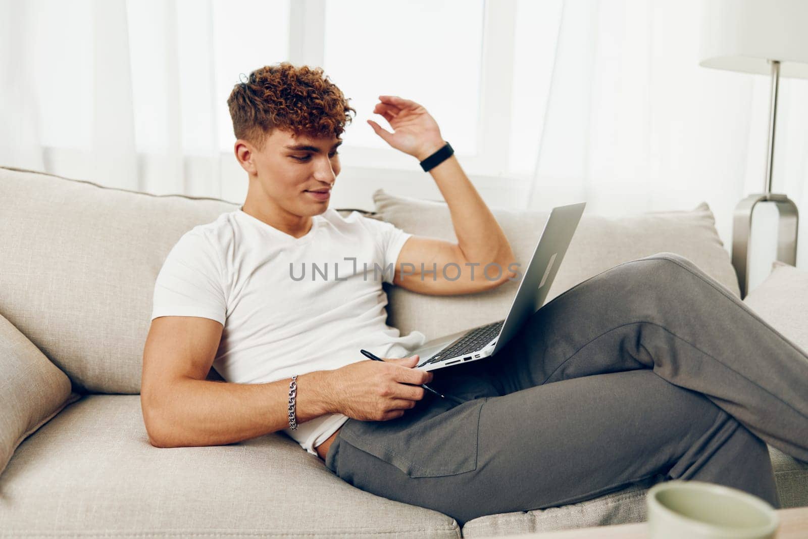 man interior home curly couch person white blissful sitting job laptop typing cup internet phone freelance adult communication browsing caucasian technology t-shirt business