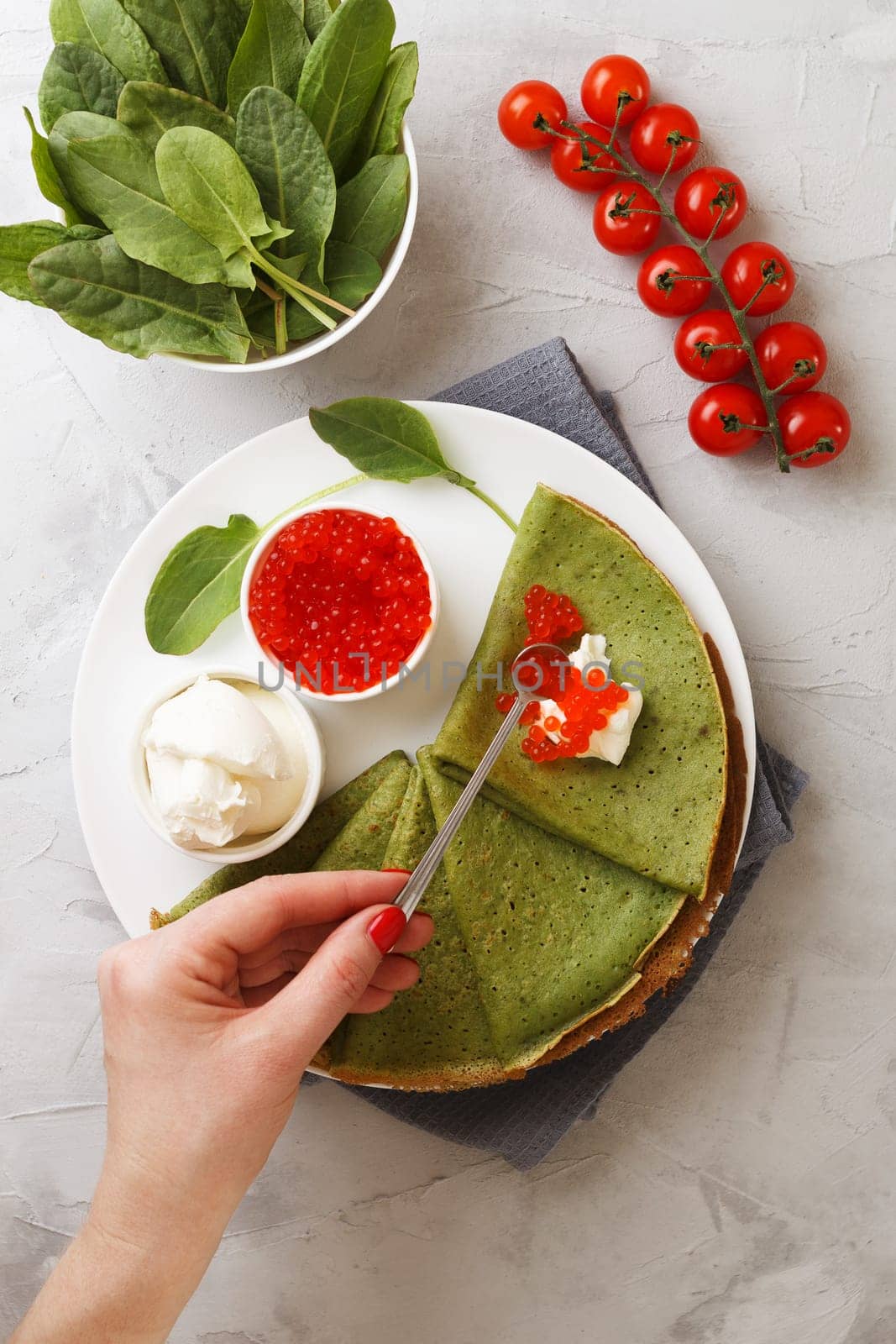 A woman lays caviar on a spinach pancake with soft cheese and cherry tomatoes on a gray background with textiles. vertical photo