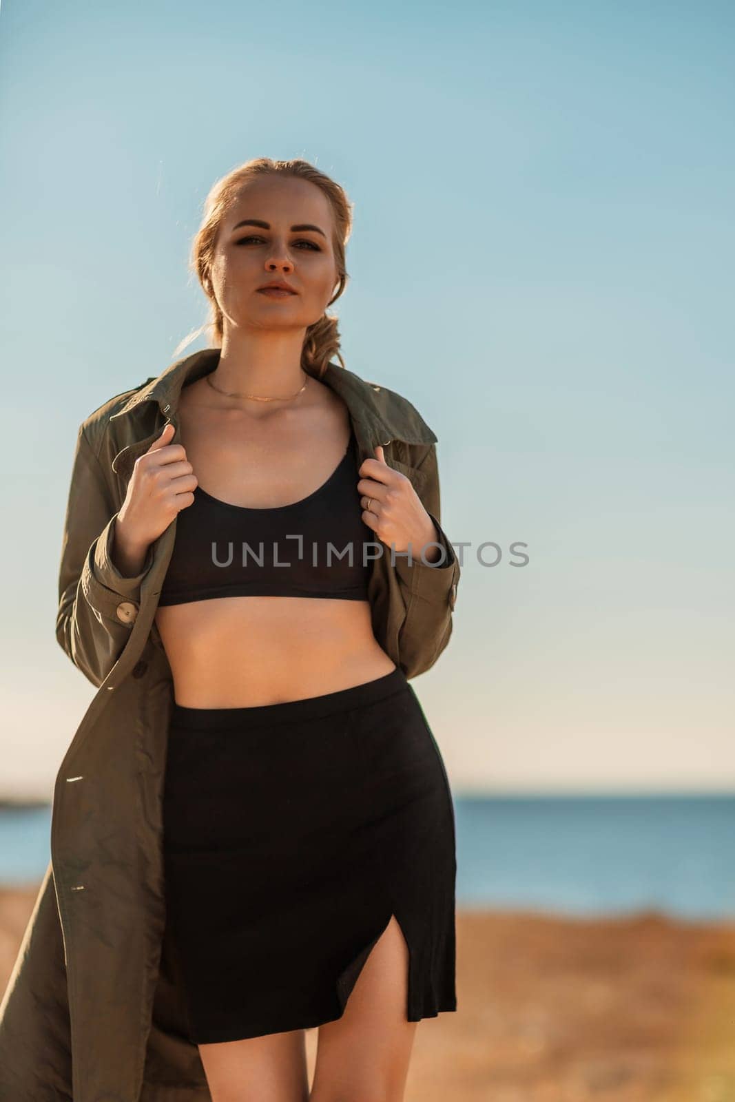 Portrait blonde sea cape. A calm young blonde in an unbuttoned khaki raincoat walks along the seashore, under a raincoat a black skirt and top by Matiunina