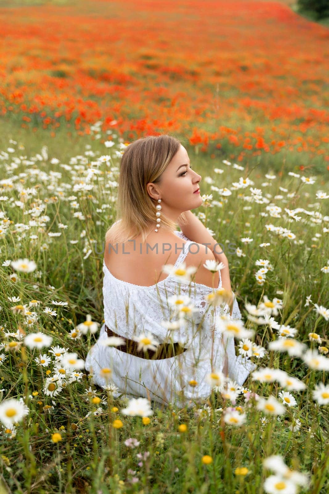 A woman sits in a chamomile field, dressed in a white dress.