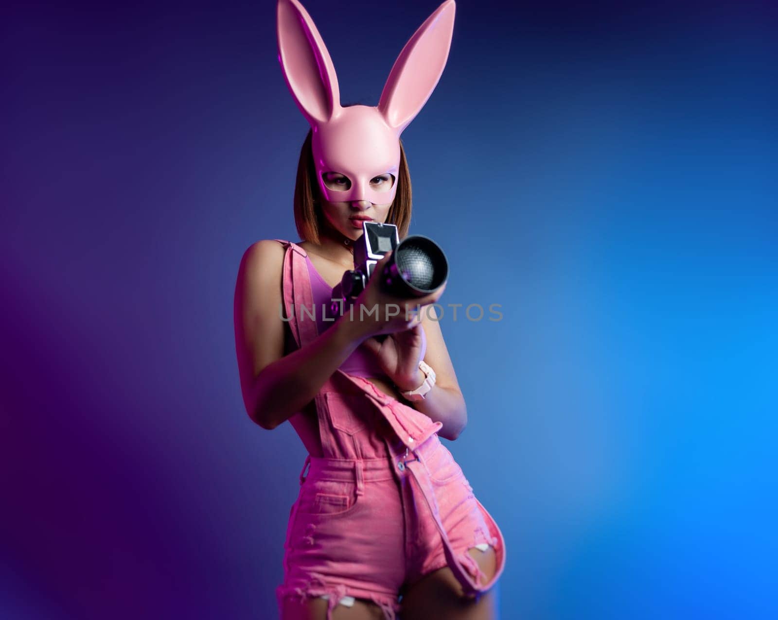 sexy girl in a pink bunny mask with a retro video camera in neon light on the background