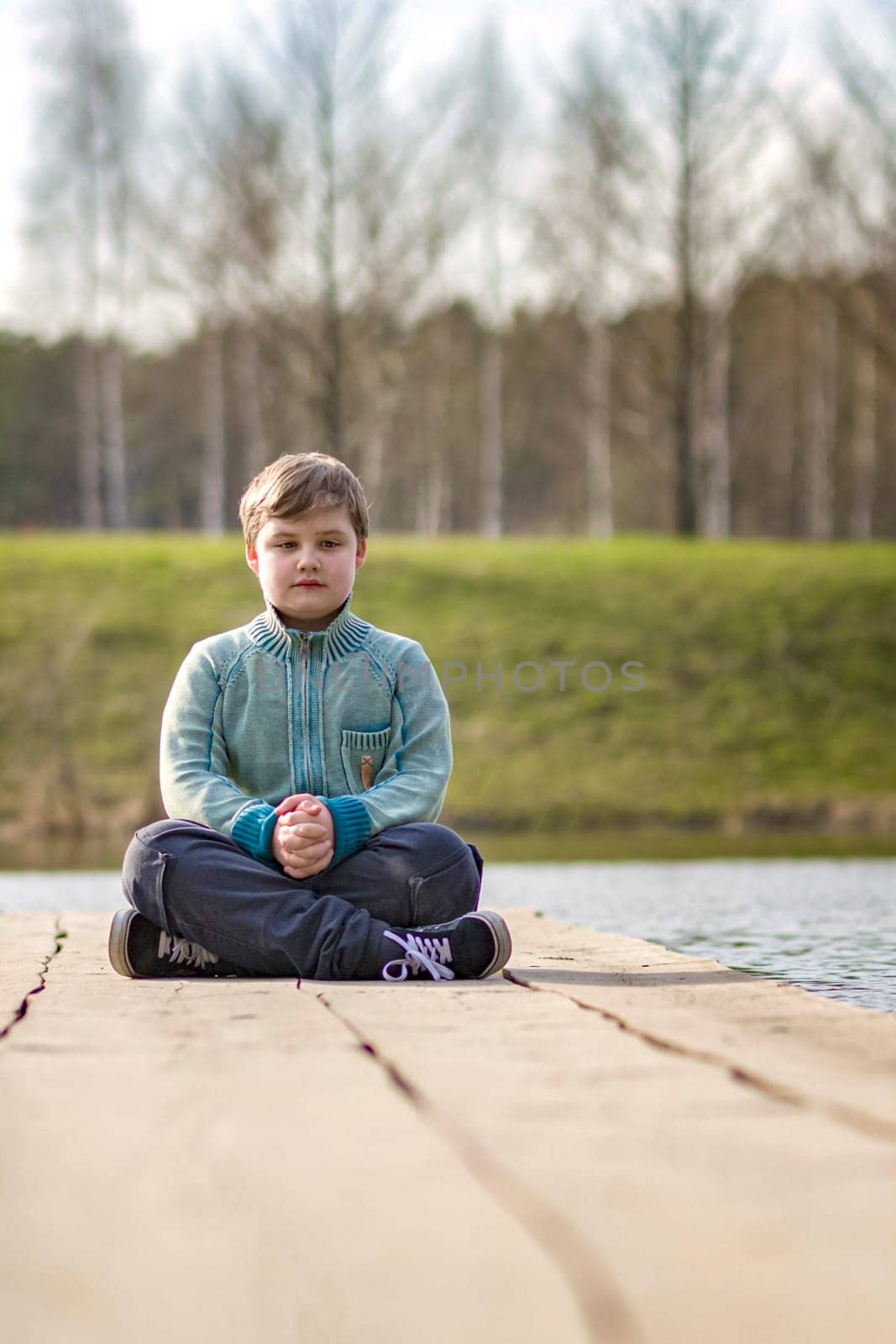 A boy is sitting on a bridge in a green park. The path is a bridge over the lake. Long wooden flooring