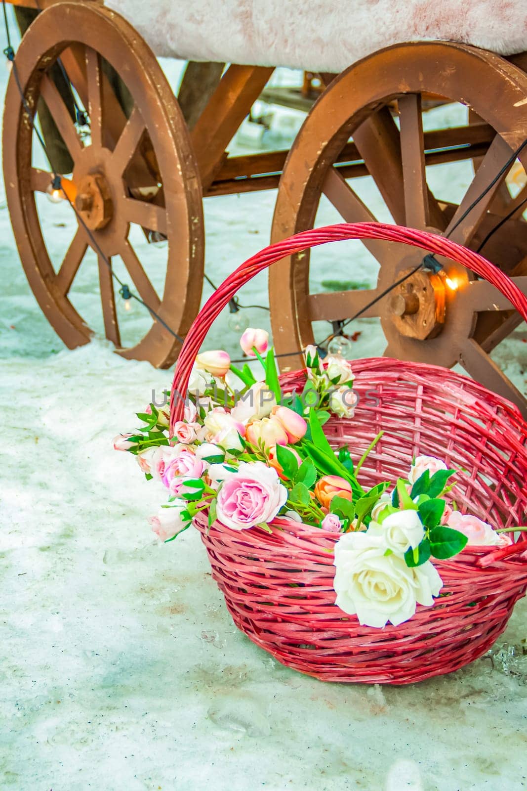 A wicker basket with beautiful flowers stands on the snow near the cart wheel. Flowers in a pink basket
