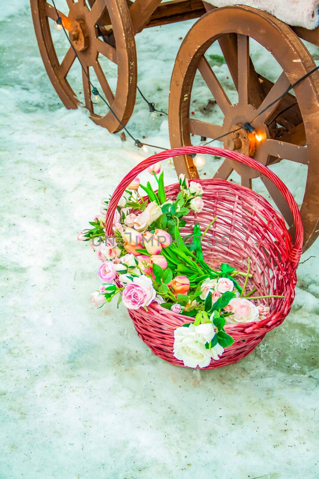 A wicker basket with beautiful flowers stands on the snow near the cart wheel. by Alina_Lebed