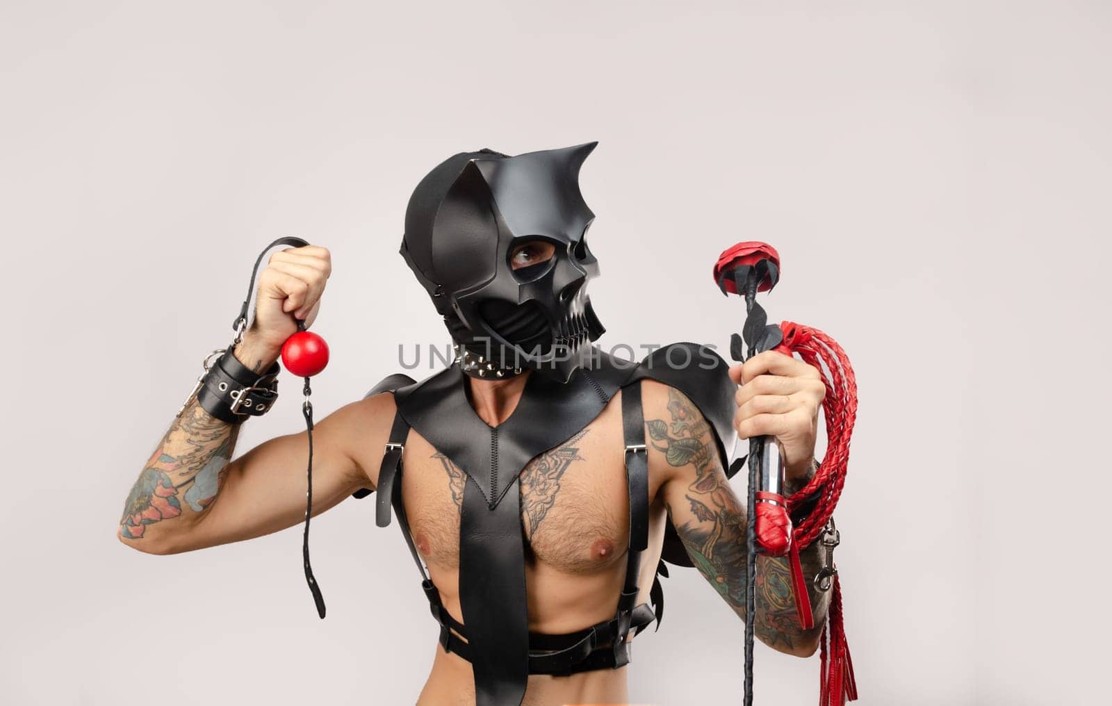 A sexy man in bdsm wearing a demon mask with a whip wearing a leather cloak with leather handcuffs and partuppets on a white background