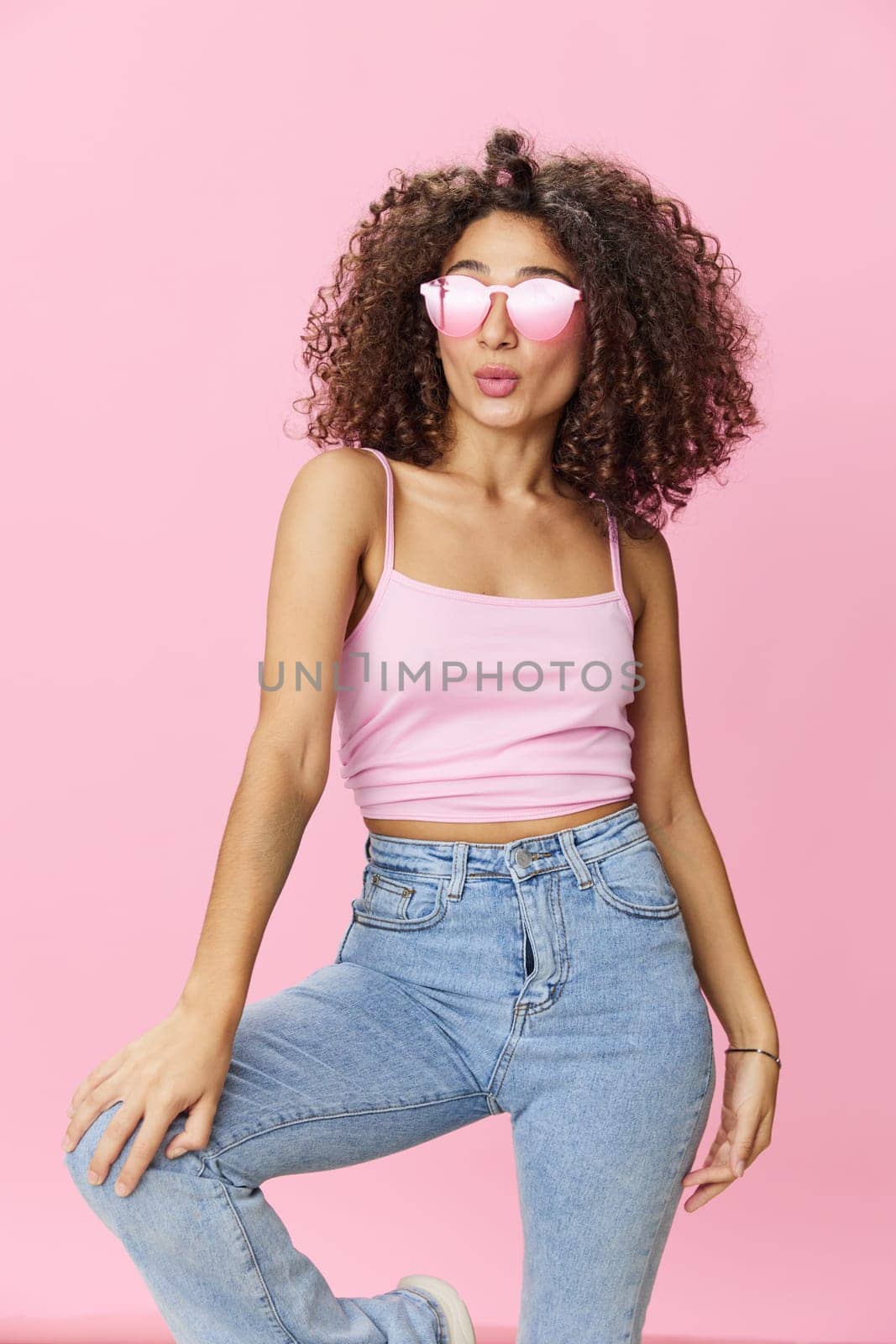Happy woman afro curls hair dancing on a pink background in summer pink t-shirt jeans and glasses, summer vibe, copy space by SHOTPRIME