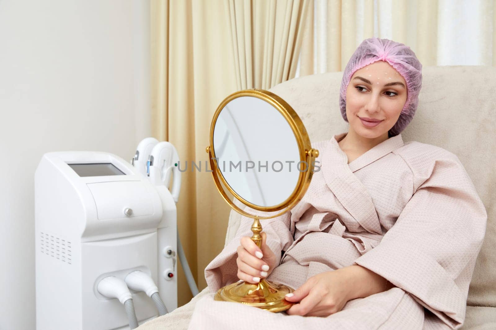 Young woman sitting in medical chair and looking in the mirror. She is satisfied after successful beauty treatment by Mariakray