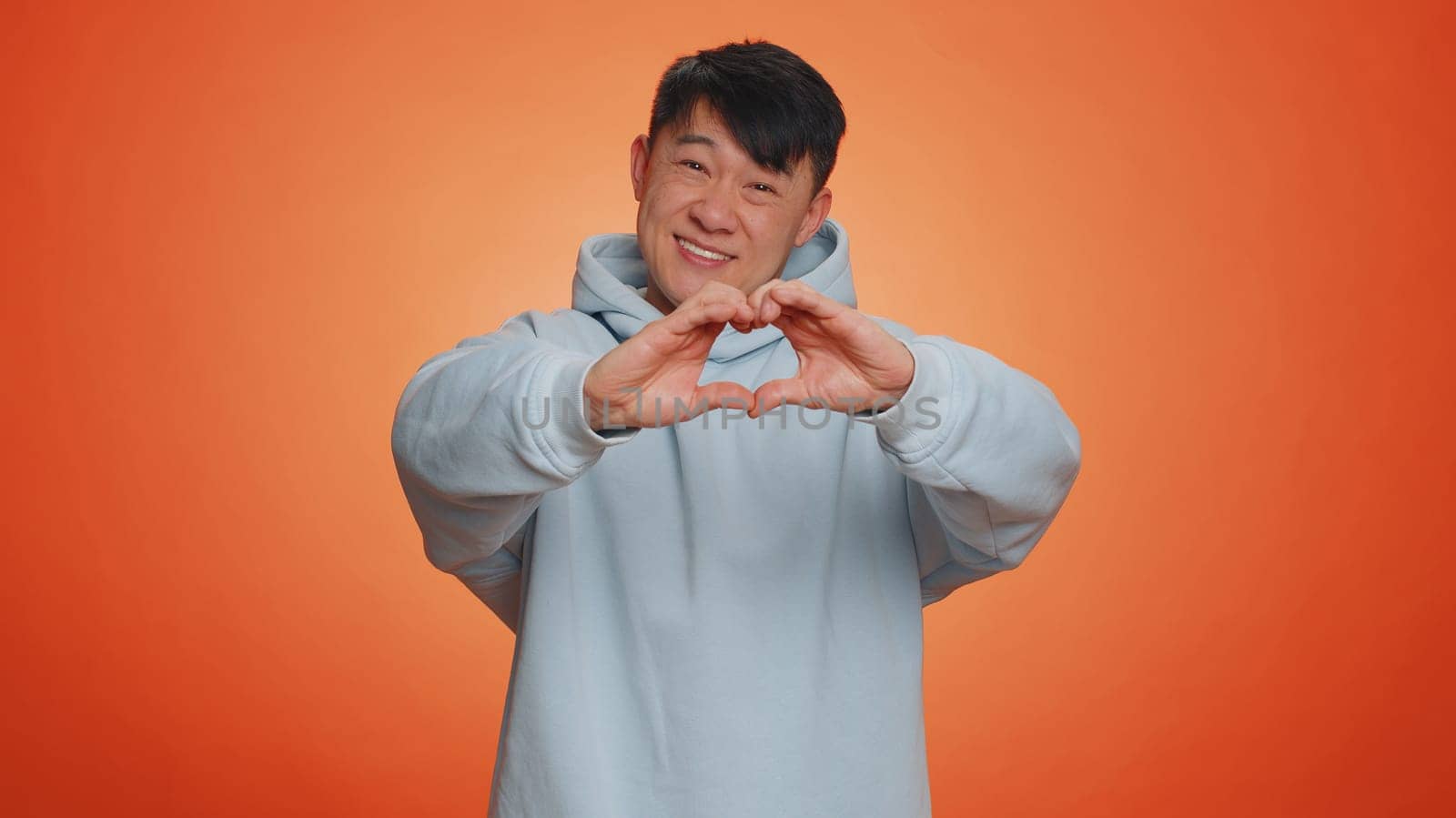 Man in love. Smiling asian man 40 years old makes heart gesture demonstrates love sign expresses good feelings and sympathy. Handsome chinese adult guy isolated alone on orange studio background