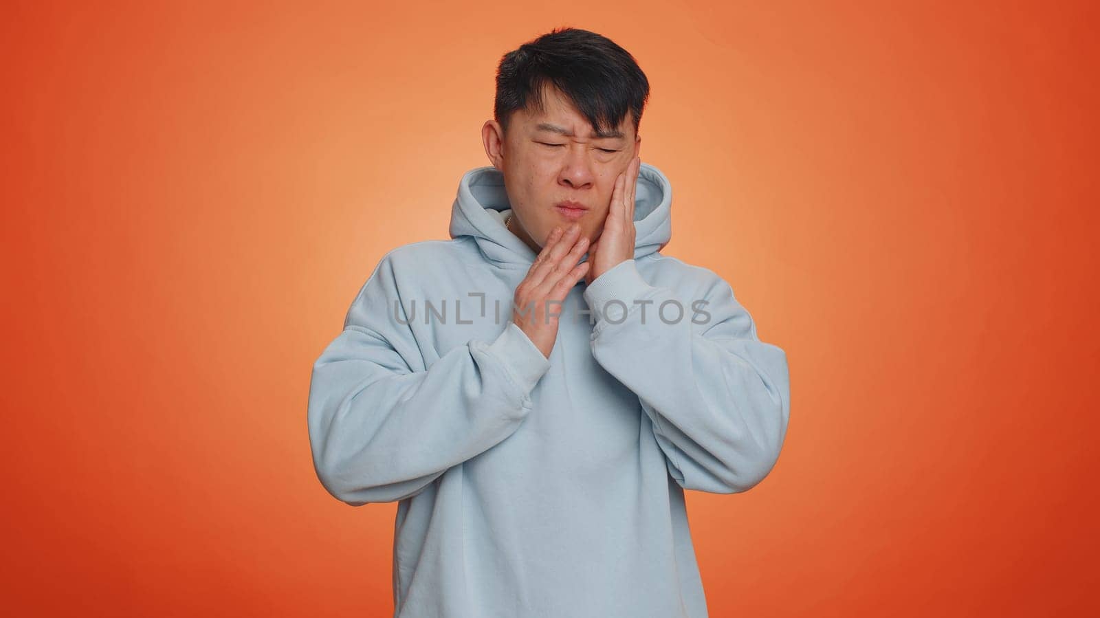 Asian man touching sore cheek suffering from toothache cavities or gingivitis waiting for dentist appointment gums disease. Handsome chinese guy indoors studio shot isolated on orange background