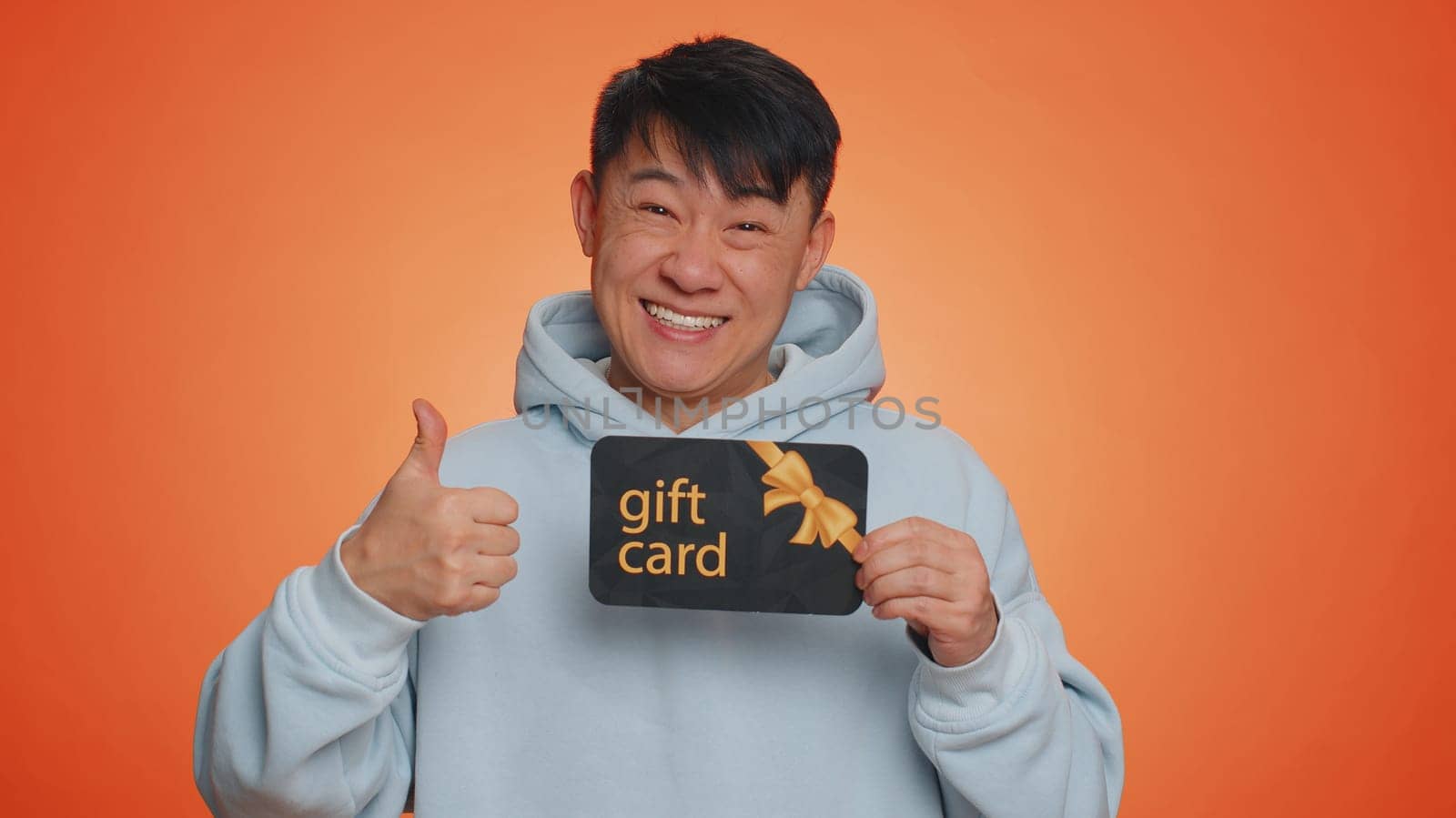 Handsome chinese man showing pointing on gift discount certificate voucher coupon for store holidays sale. Asian guy with shopping christmas surprise card isolated alone on orange studio background