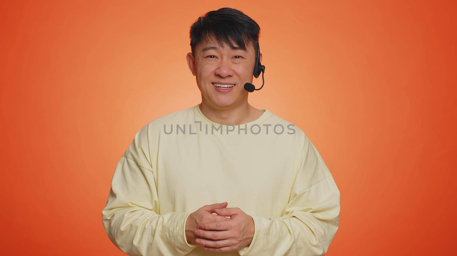 Smiling asian man wearing headset, freelance worker, call center or support service operator helpline, having talk with client or colleague, communication support. Handsome guy on orange background
