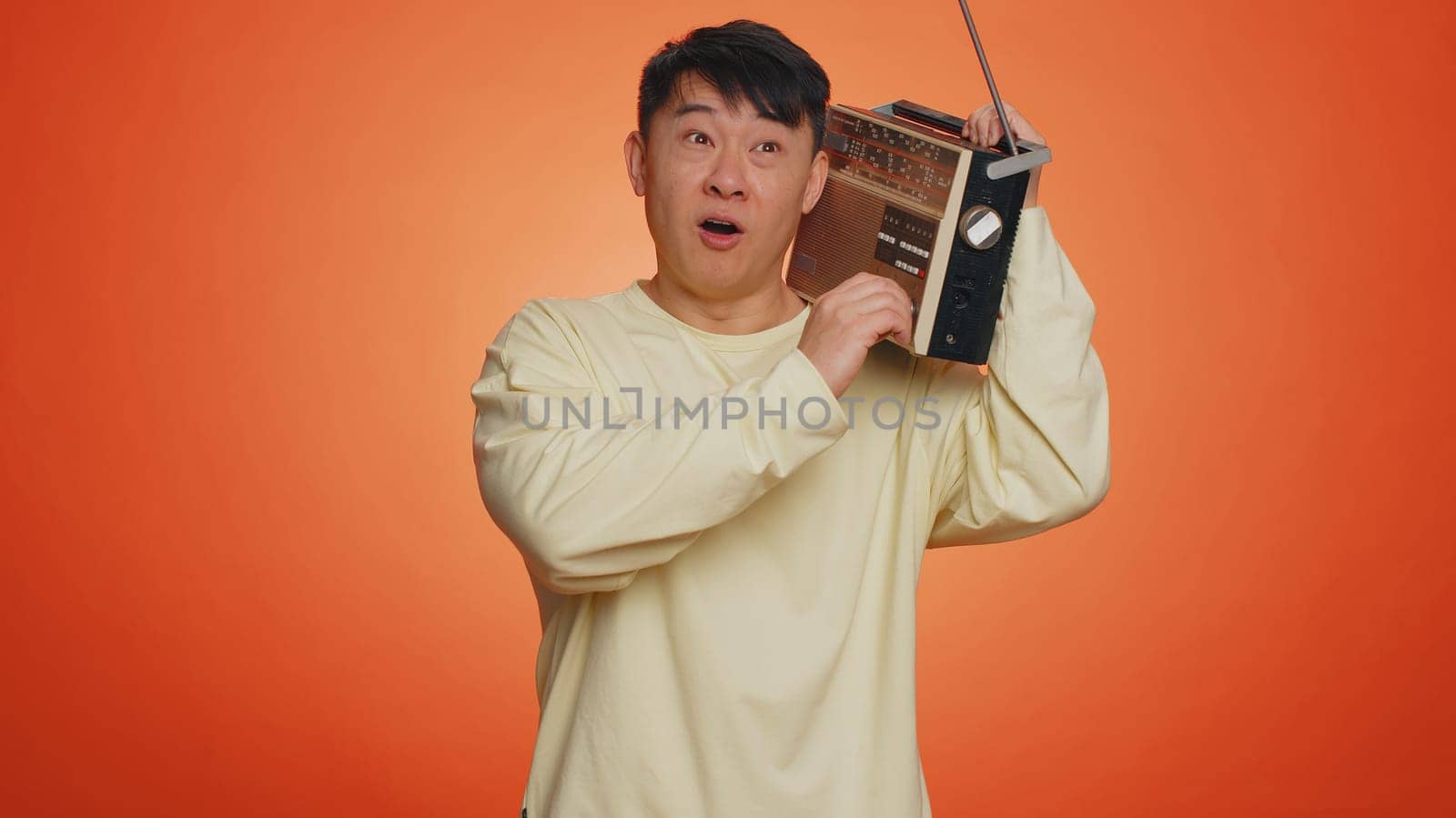 Asian man using retro tape record player to listen music, disco dancing of favorite track, having fun, entertaining, fan of vintage technologies. Handsome chinese guy isolated on orange background