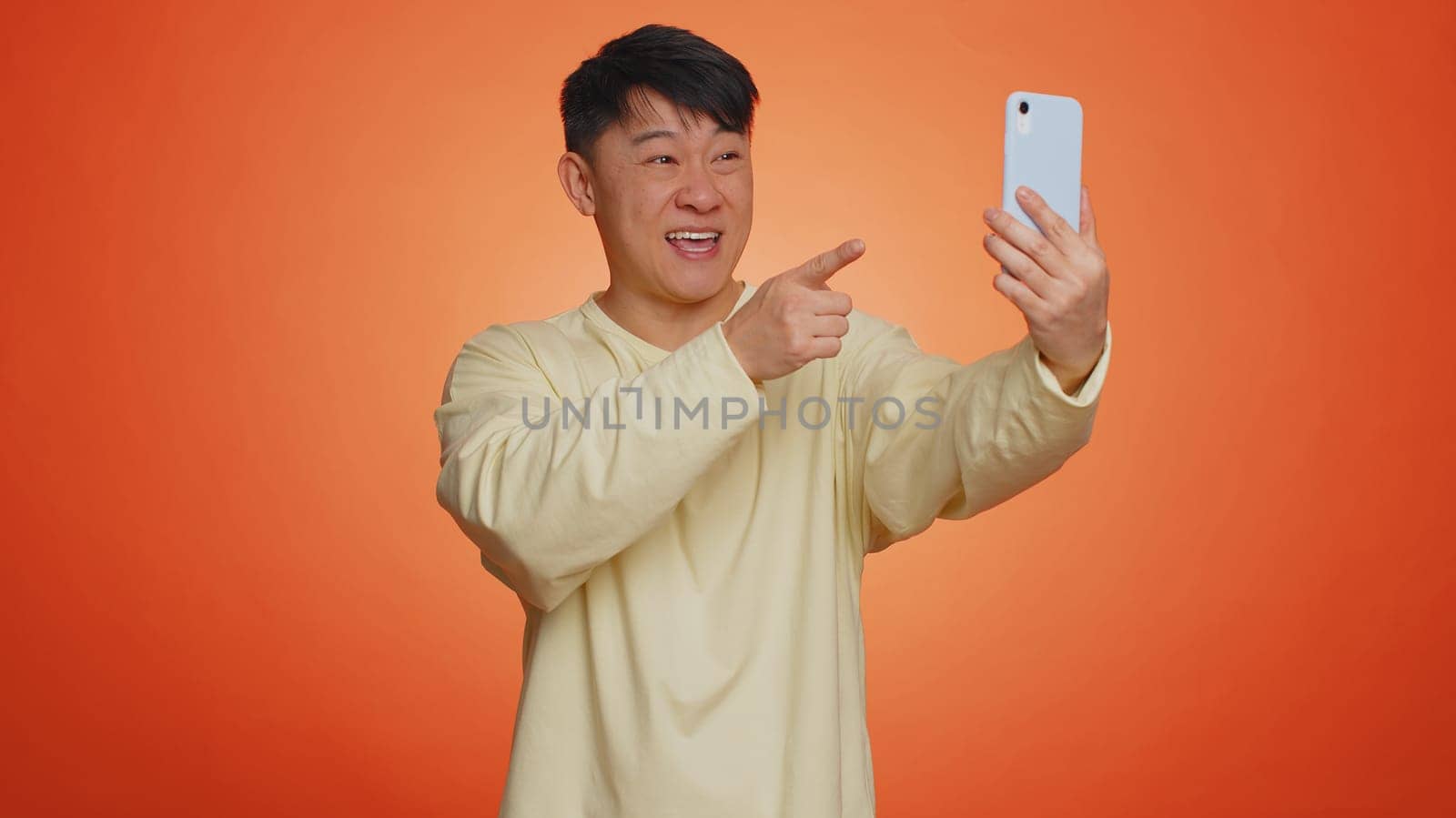 Asian man traveler blogger, taking selfie on smartphone, communicating video call online with family or friends, live translation stream. Handsome chinese guy isolated on orange background, indoors